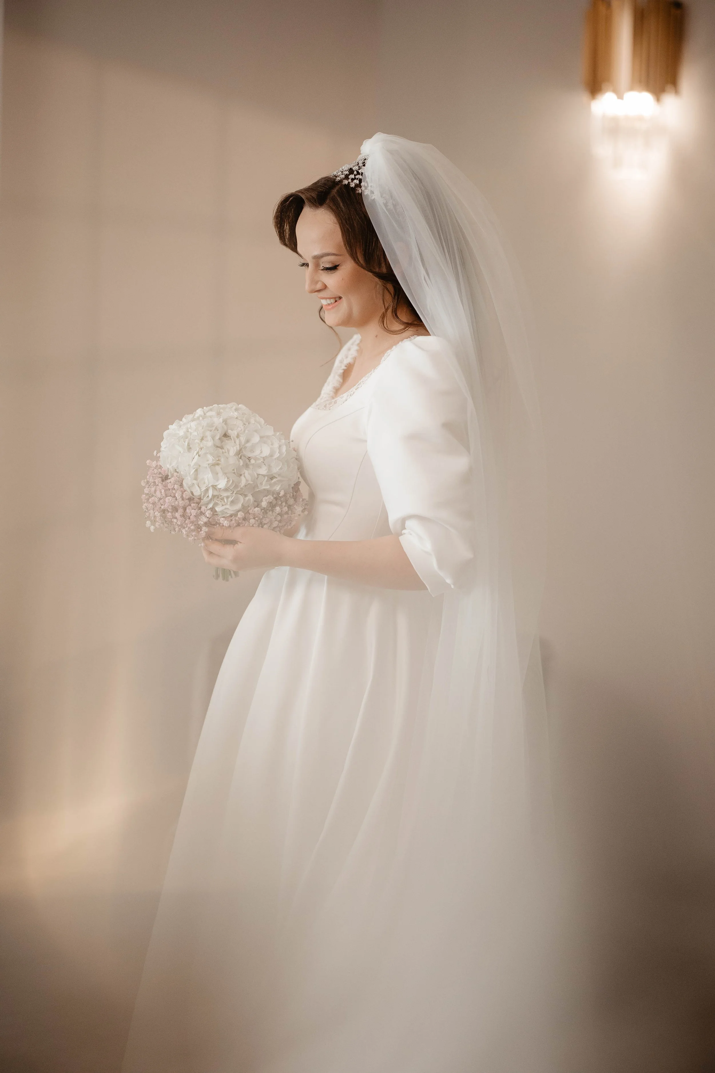A bride in a white wedding dress with puffed sleeves and a lace neckline, holding a bouquet of white and blush pink flowers, standing indoors with beige walls and warm lighting.