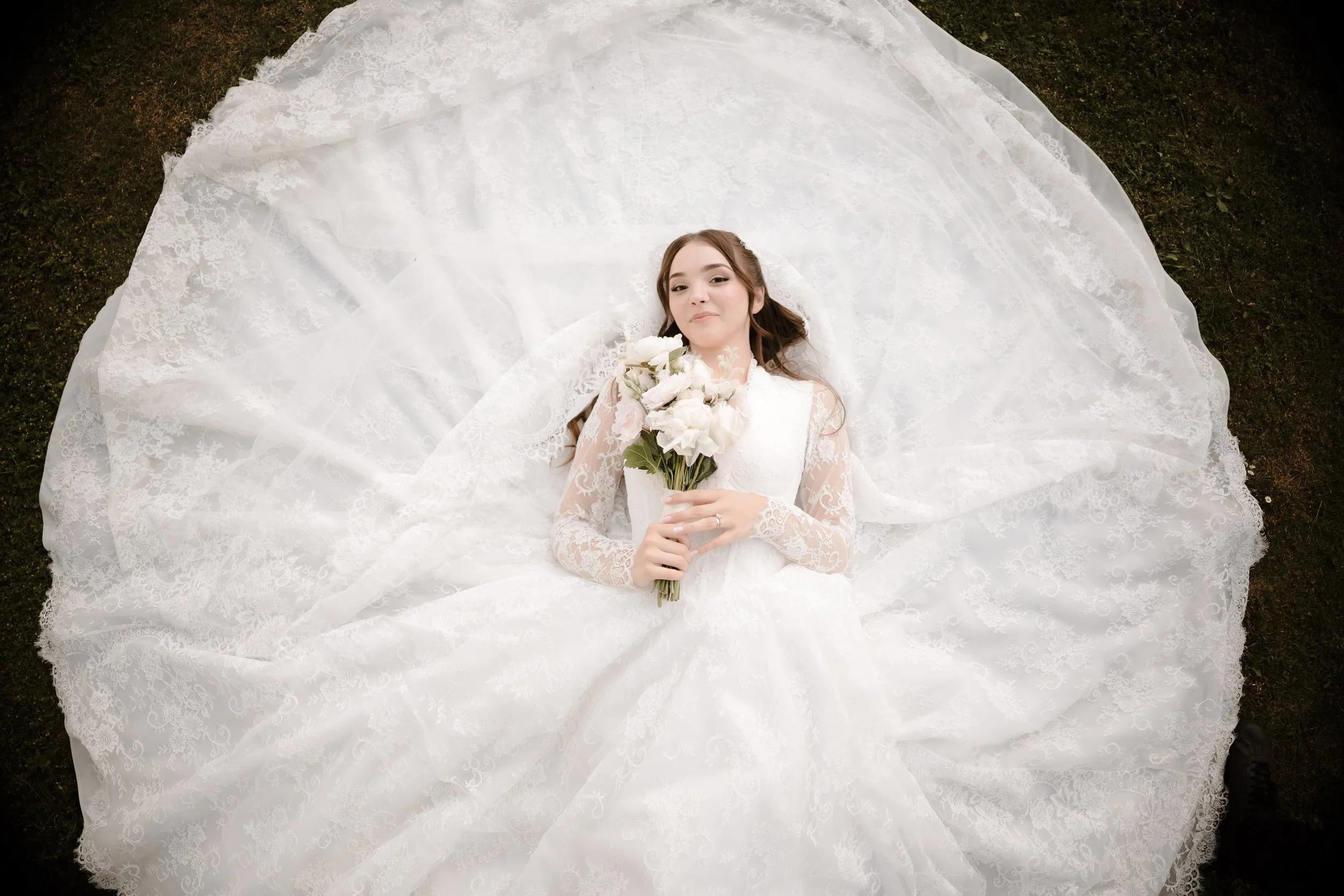 A bride lying on the ground on her back in a wedding dress and holding a bouquet of white flowers, with her head towards the camera and her dress spread out around her.