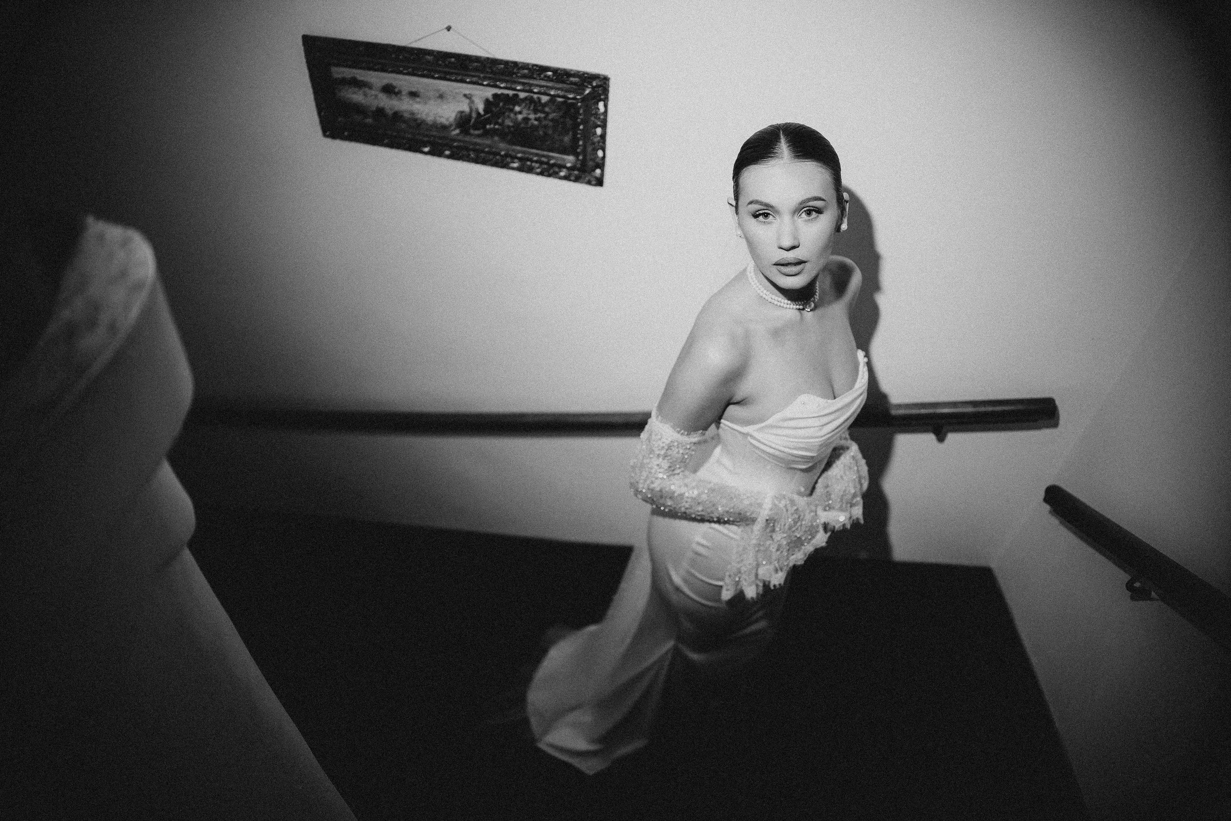 Black and white photo of a woman with dark hair slicked back, wearing an off-the-shoulder dress and jewelry, standing in a room with plain walls. The woman looks directly at the camera, while a person, partially visible on the left, appears to be tak