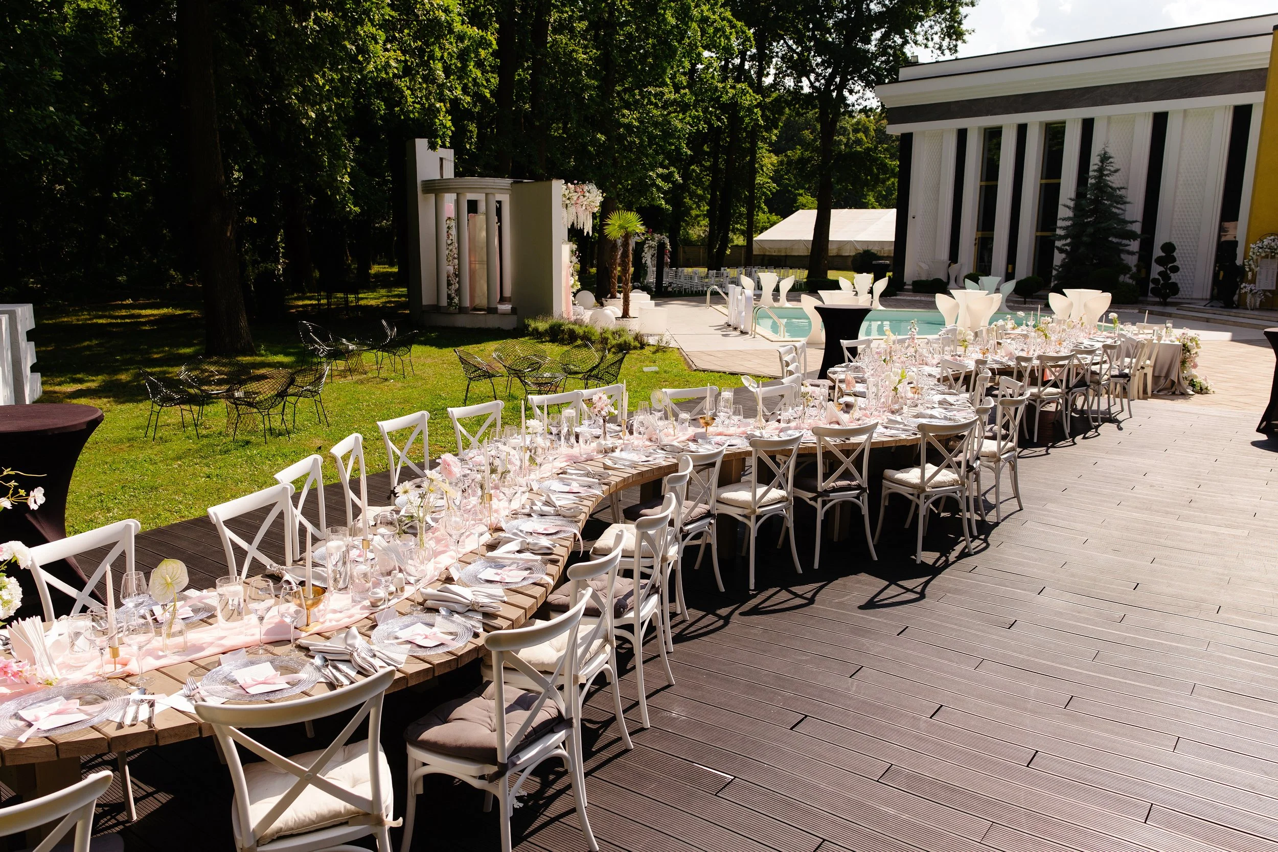 Outdoor event setup with round and rectangular tables, white chairs, and floral decorations on a wooden deck near a pool, with trees and a building in the background.