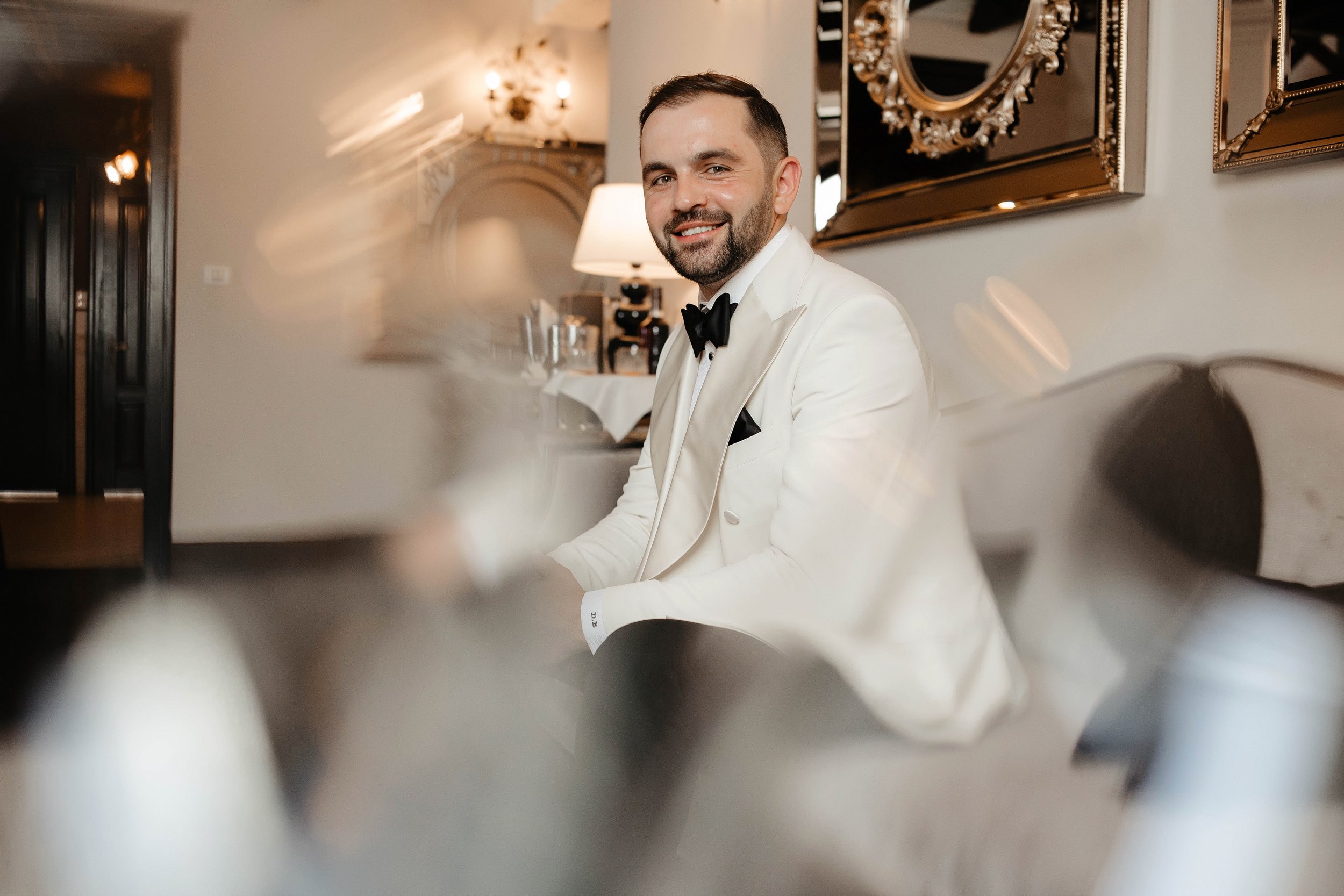 A man dressed in a white tuxedo with a black bow tie, smiling, sitting in a decorated room with elegant mirrors and warm lighting.