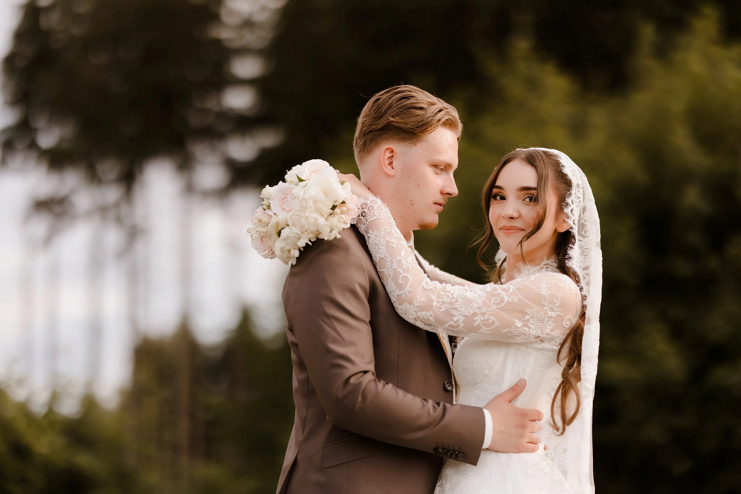 A young bride and groom are embracing outdoors, with the groom holding a bouquet of white and pink roses. The bride is wearing a lace wedding dress and veil, and they are surrounded by greenery and trees.
