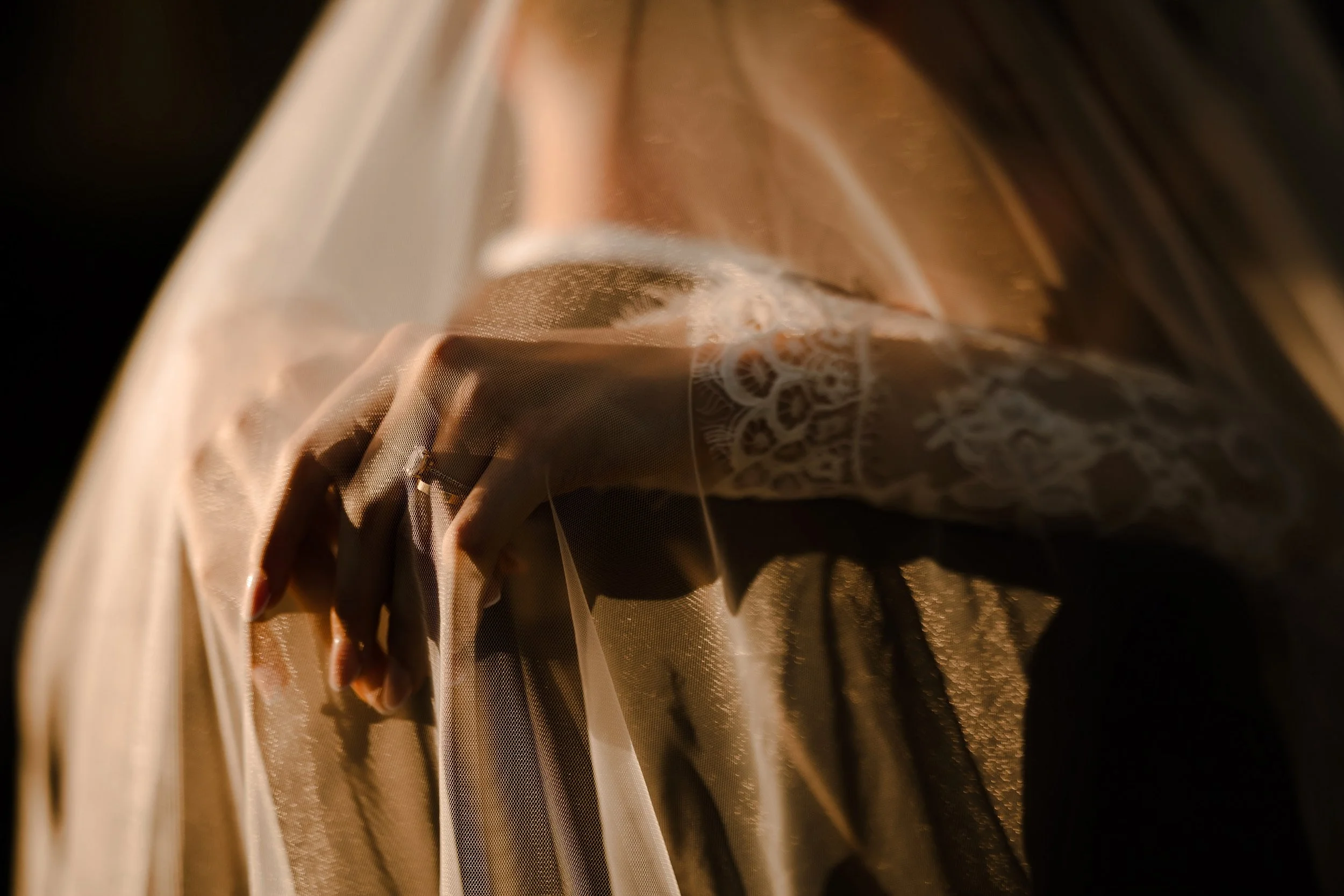Close-up of a hand holding a piece of delicate, sheer fabric with intricate lace or embroidery details, illuminated with warm, soft lighting.