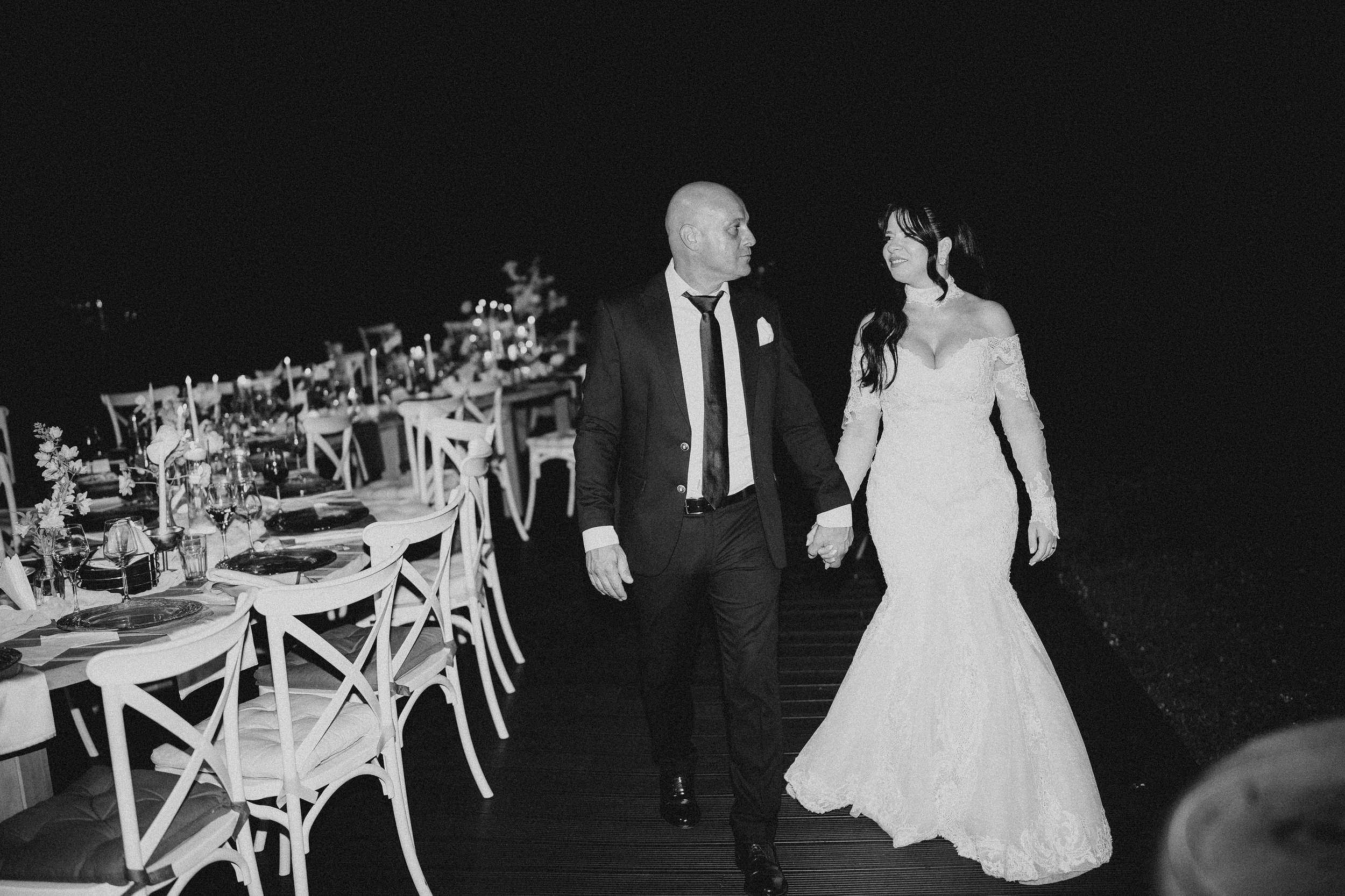 A black and white photo of a bride and an older man, possibly her father, walking hand-in-hand at a wedding reception. The bride is wearing a lace off-the-shoulder wedding gown, and the man is dressed in a suit with a tie. The reception area has deco