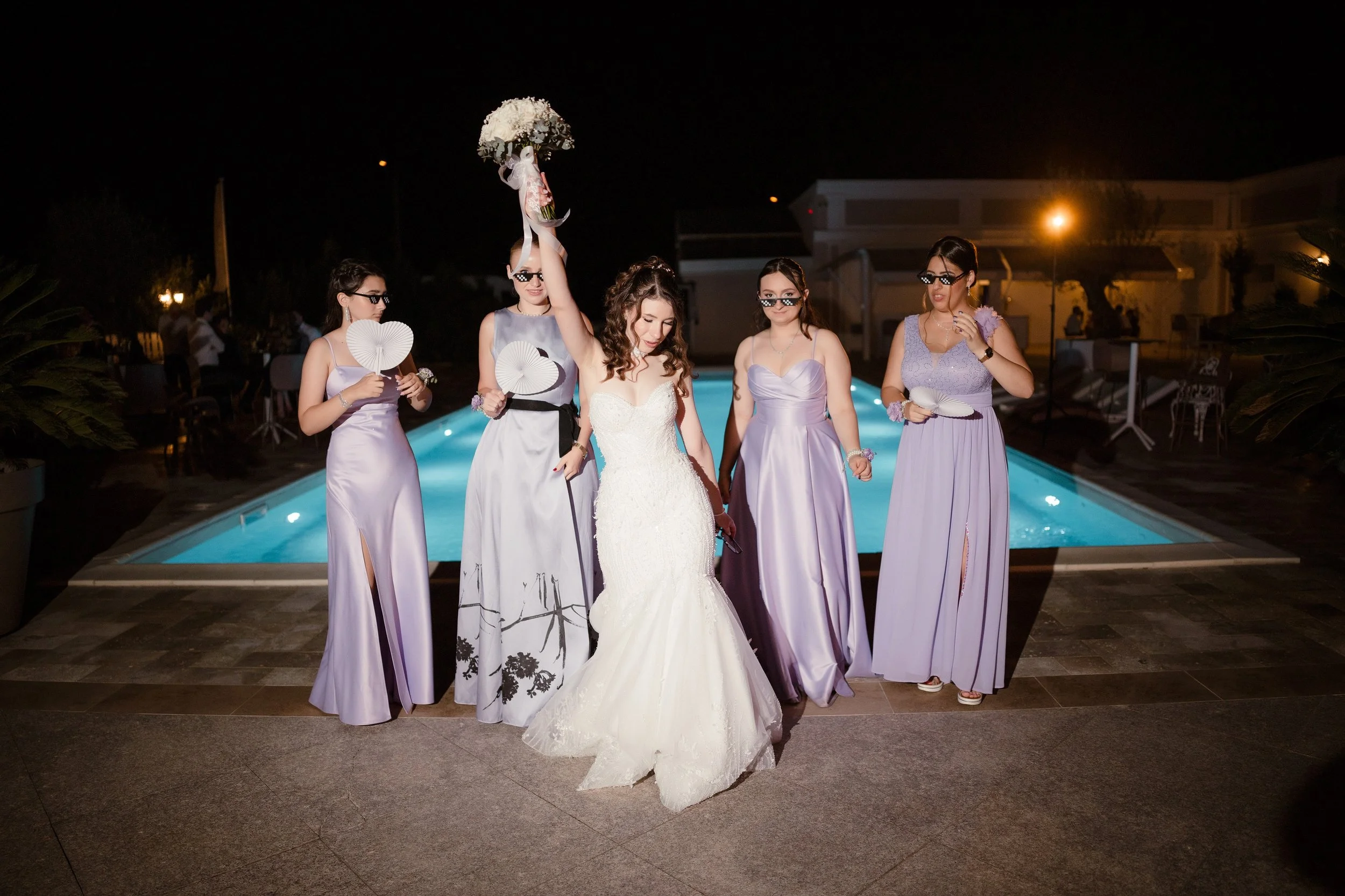 A bride in a white wedding gown holding a bouquet of flowers raised in the air, surrounded by five women in lavender dresses and black sunglasses, all standing by a swimming pool at night.