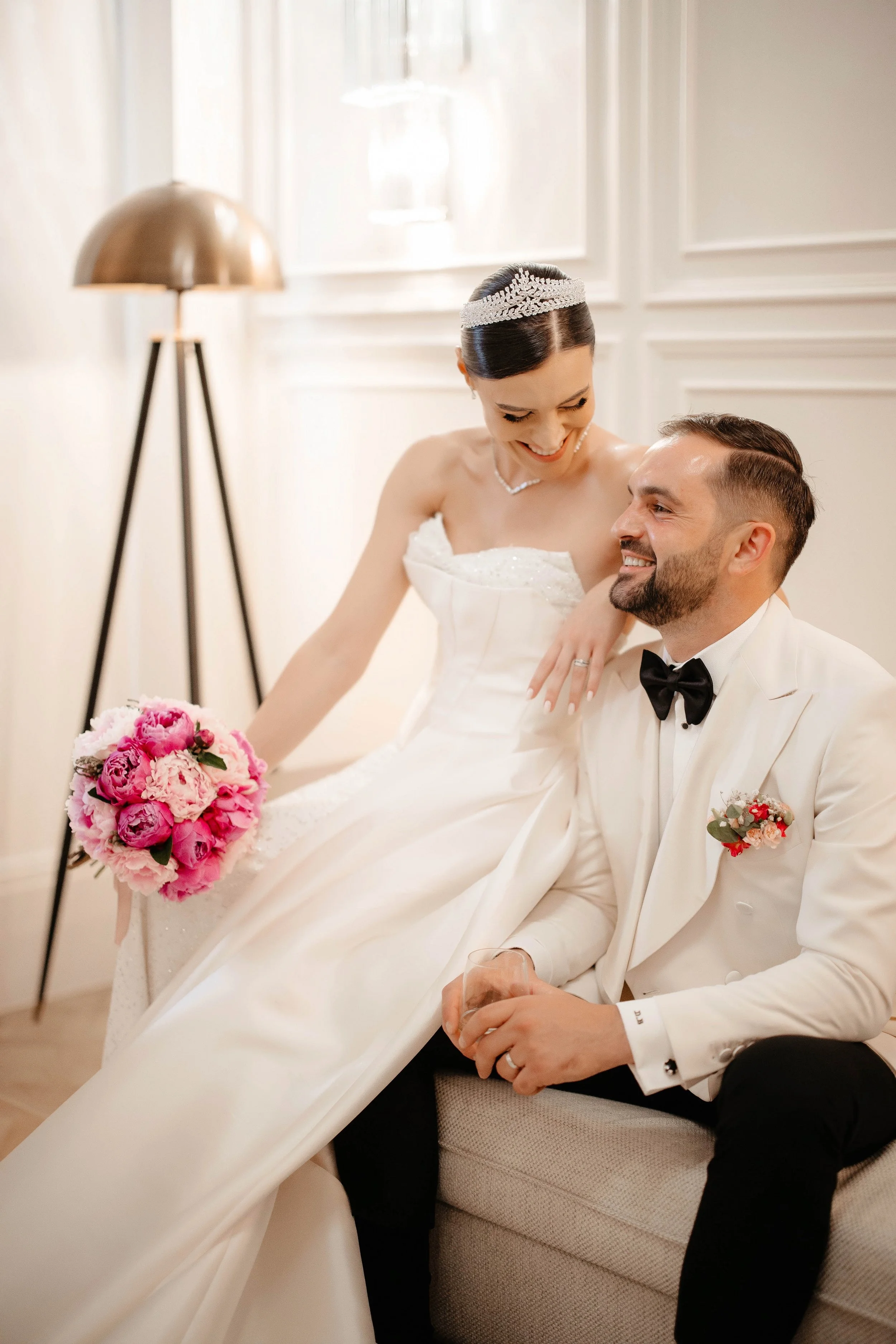 A bride and groom sitting together, smiling, with the bride holding a pink bouquet of flowers. The bride wears a white wedding dress and a tiara, and the groom wears a white tuxedo with a black bow tie and boutonniere. They are in a well-lit room wit