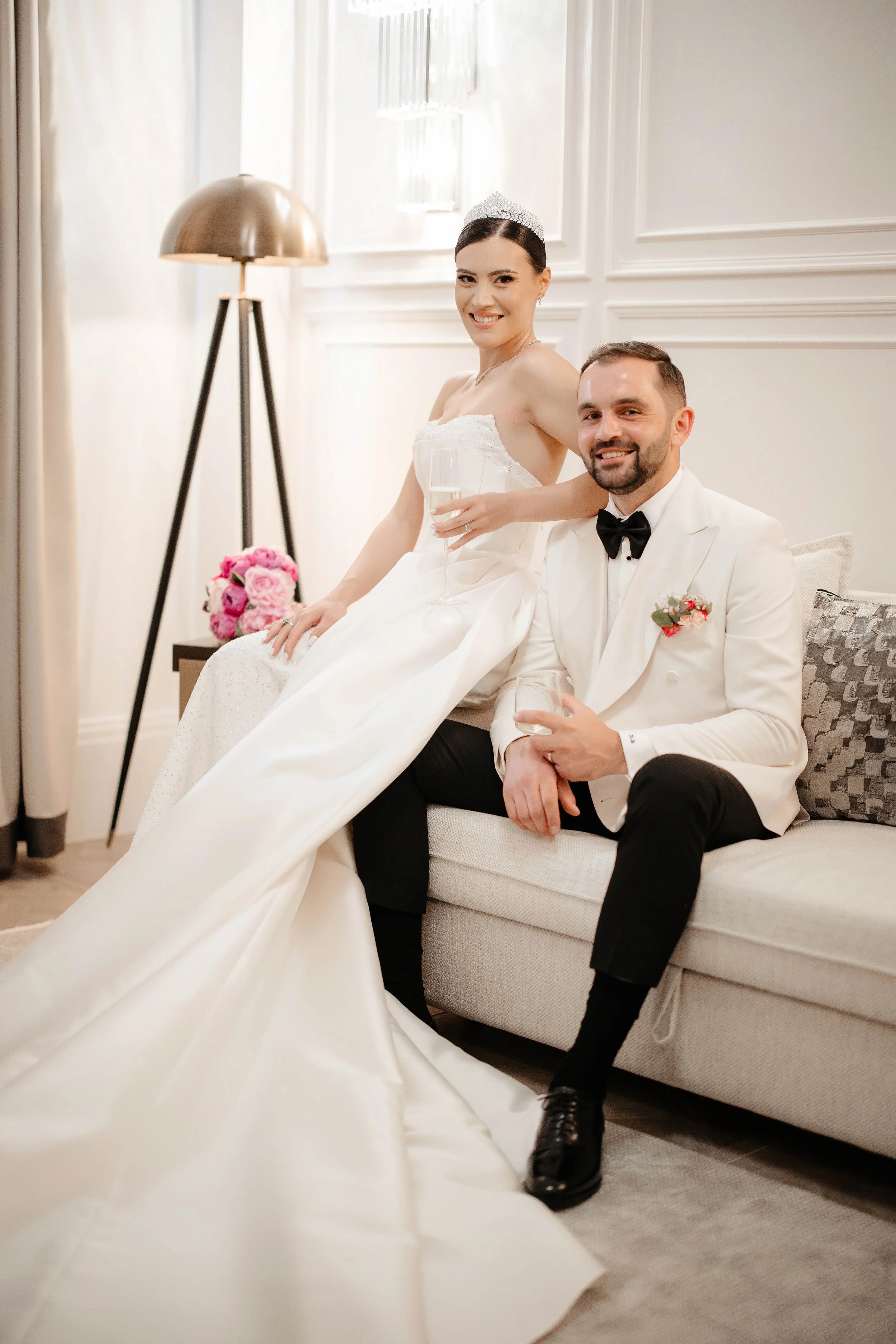 A bride and groom sitting on a white couch at their wedding reception. The bride is holding a glass of champagne and wearing a tiara, while the groom is dressed in a tuxedo with a bow tie, smiling.