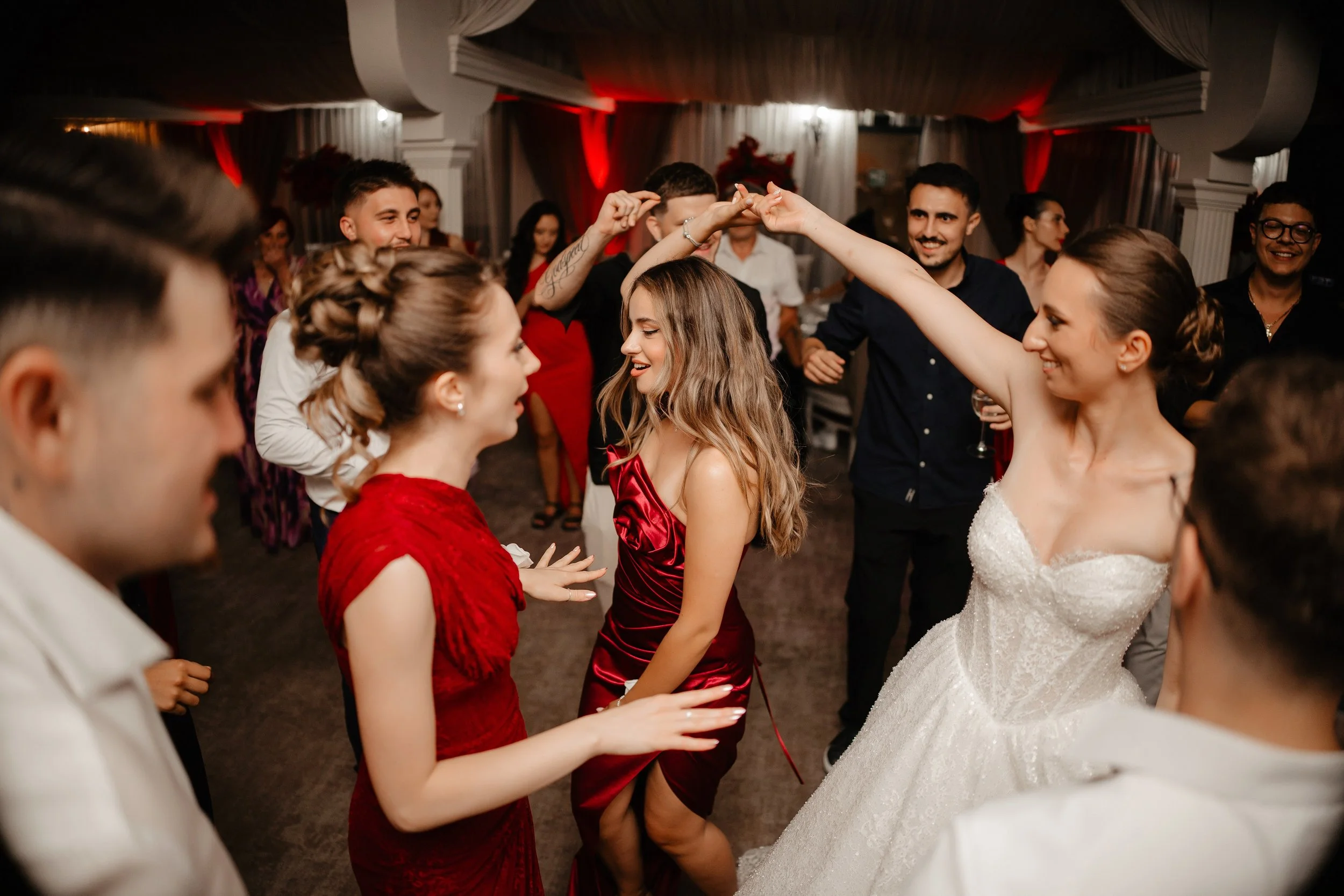 People dancing at a wedding reception, with women in red dresses and a bride in a white gown.