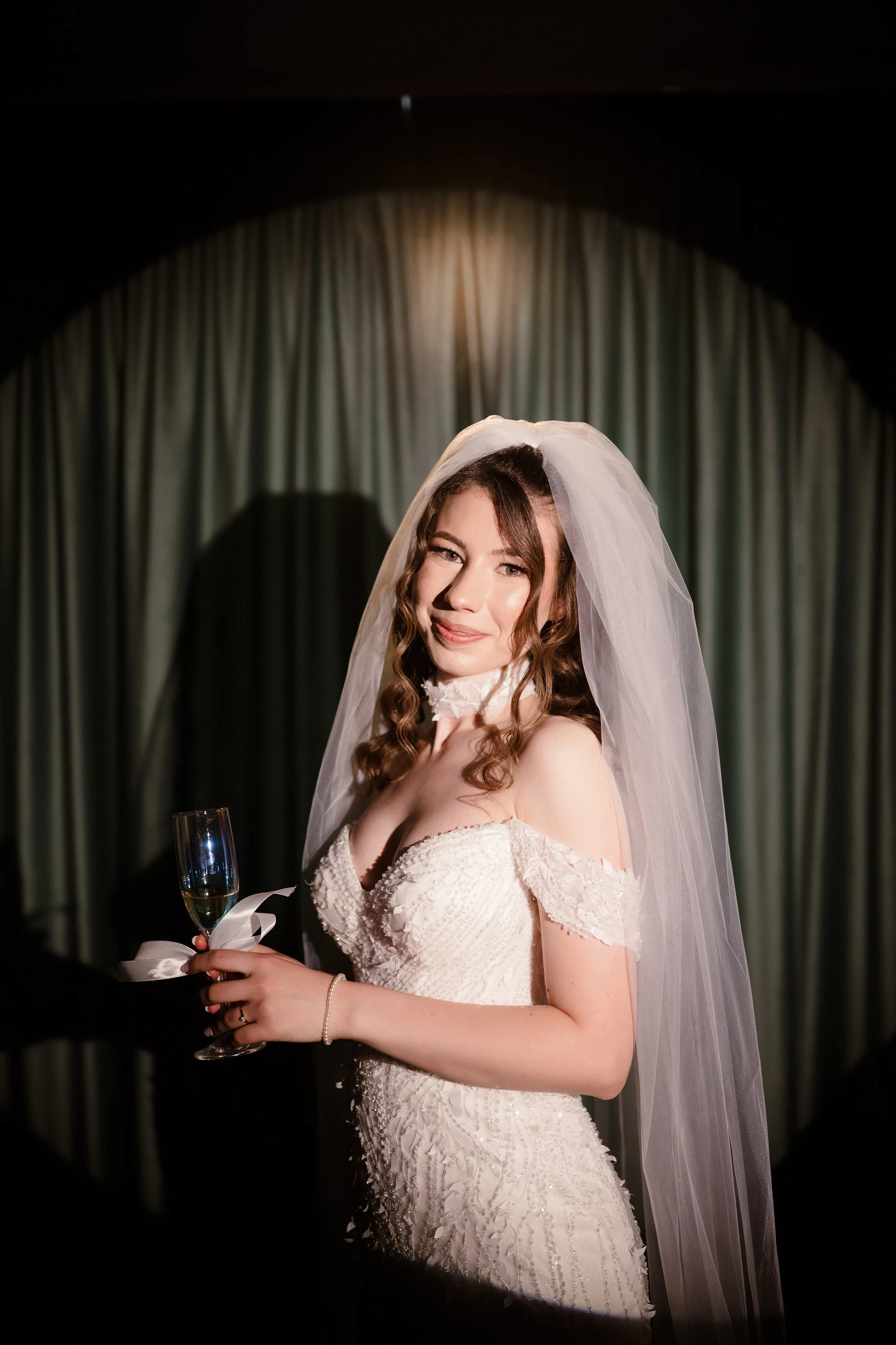 A bride with brown, curly hair in a white wedding dress with lace details, wearing a veil, holding a champagne glass, smiling softly, standing in front of dark curtains with a spotlight overhead.