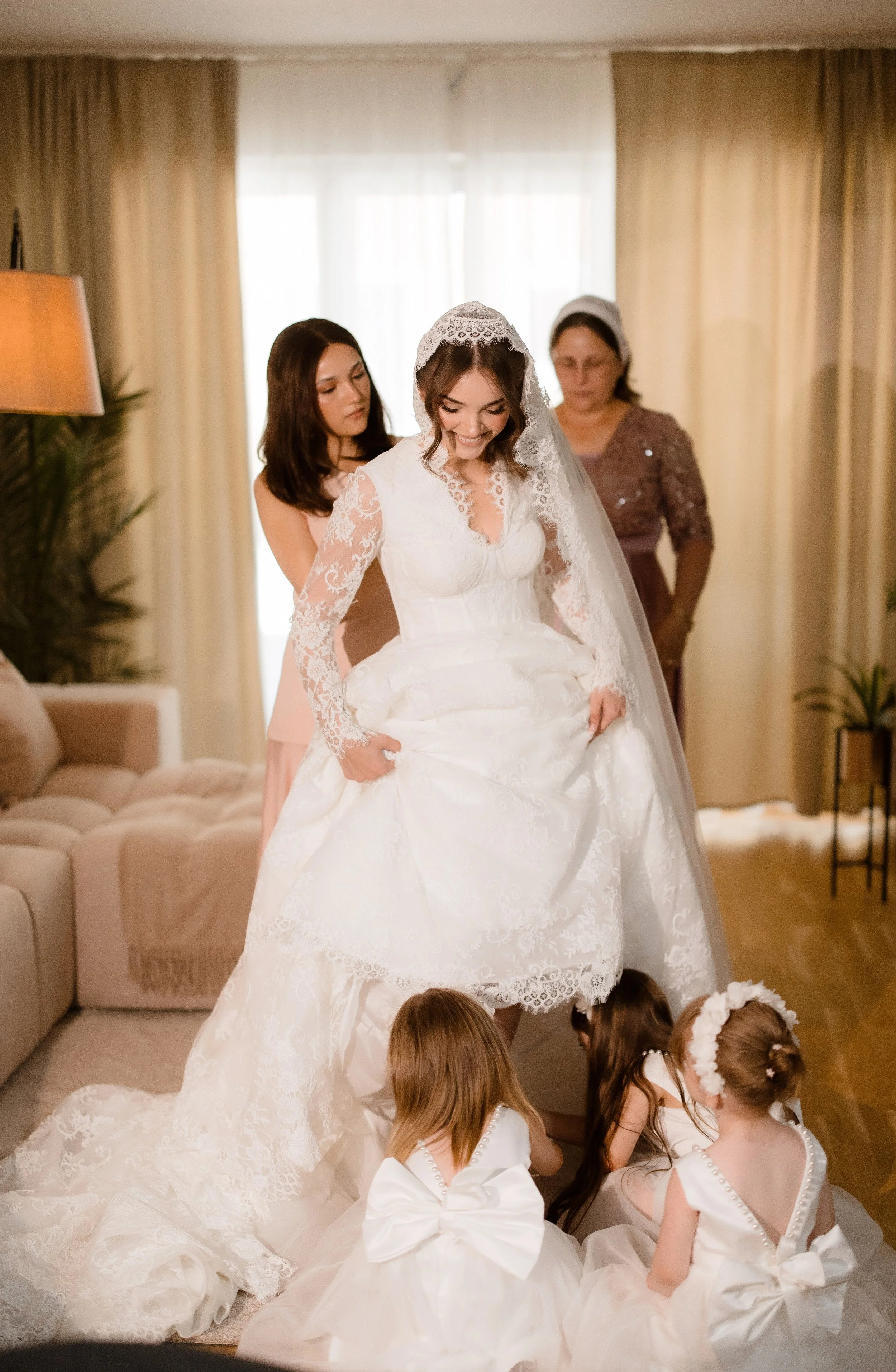 A bride in a white lace wedding dress and veil stands with her head slightly bowed, surrounded by her bridesmaids and flower girls, inside a warmly lit living room with beige curtains and a tan sofa.