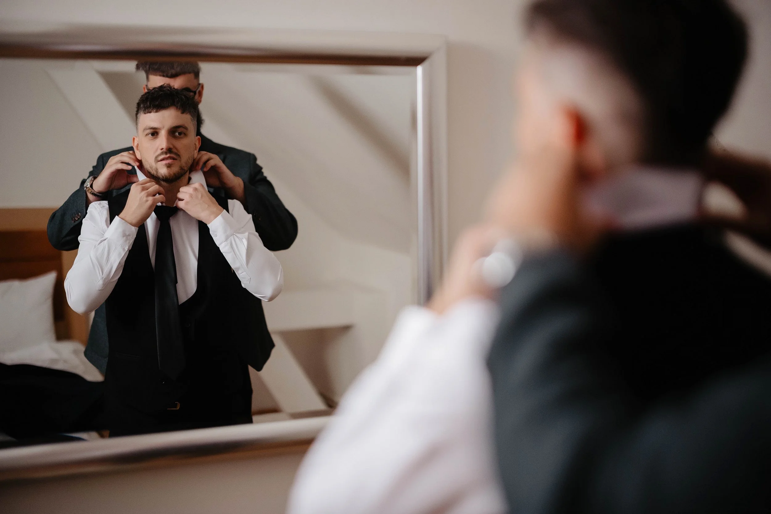 A man in a tuxedo is looking into a mirror while another person adjusts his collar from behind. The scene appears to be in a bedroom or dressing area.