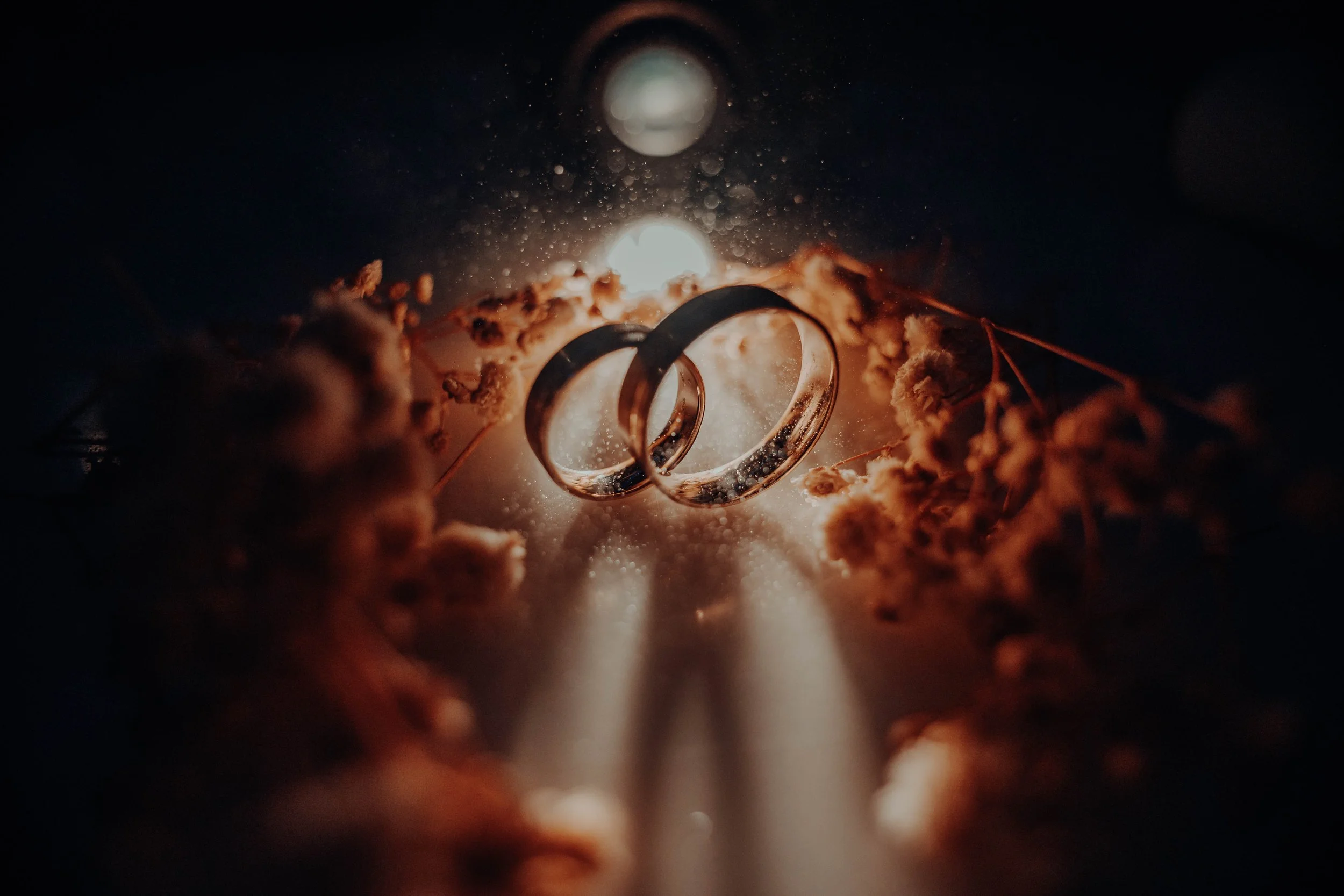 Close-up of two wedding rings suspended amid glowing sparks and dust particles with a dark background, creating a romantic and dramatic effect.