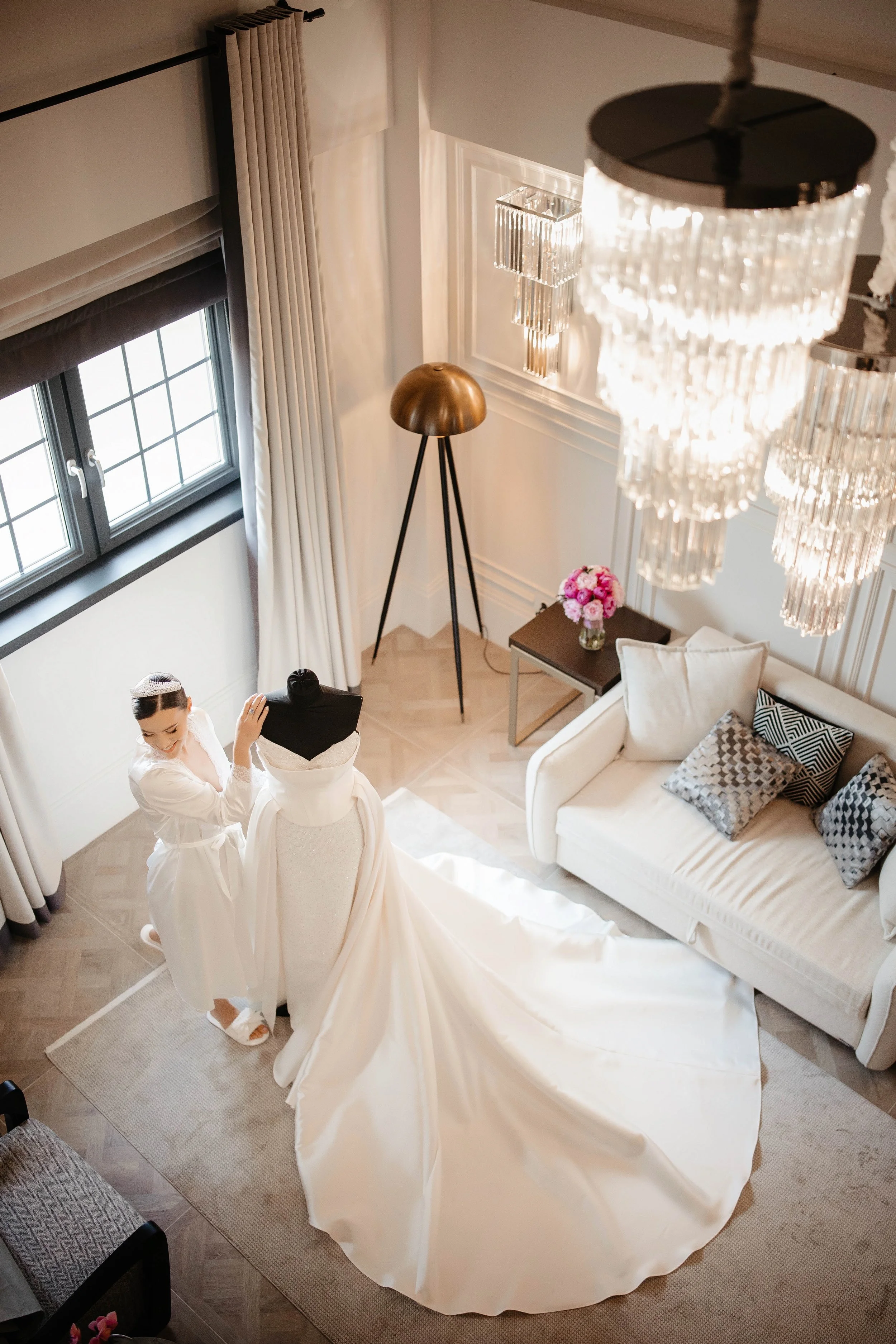 Two women in white robes preparing a wedding dress in a well-lit, elegant living room with a chandelier, a window with curtains, a bronze-colored floor lamp, a small table with pink flowers, and a white sofa with patterned pillows.