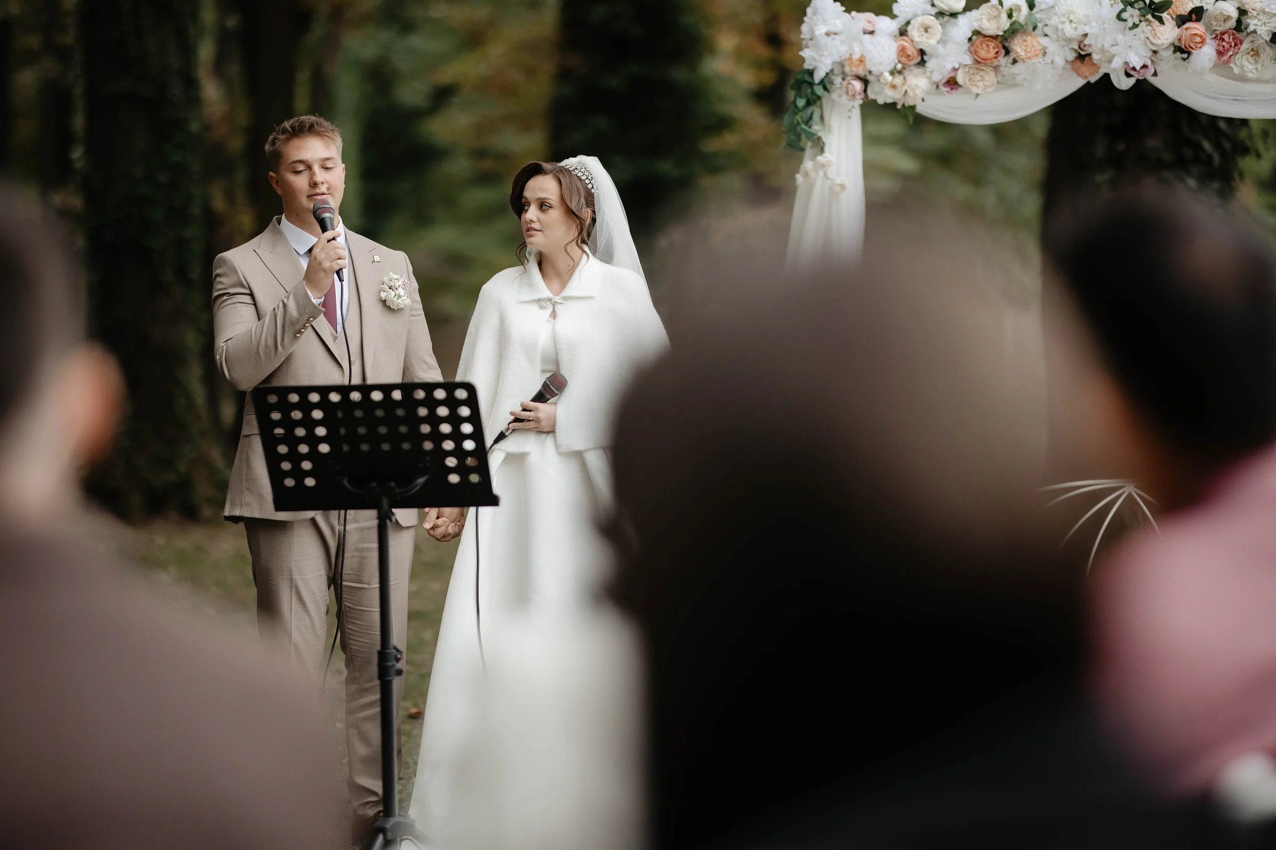A couple getting married outdoors, standing hand in hand, with a man reading vows into a microphone, and a woman dressed in white, under a floral arch, surrounded by trees.