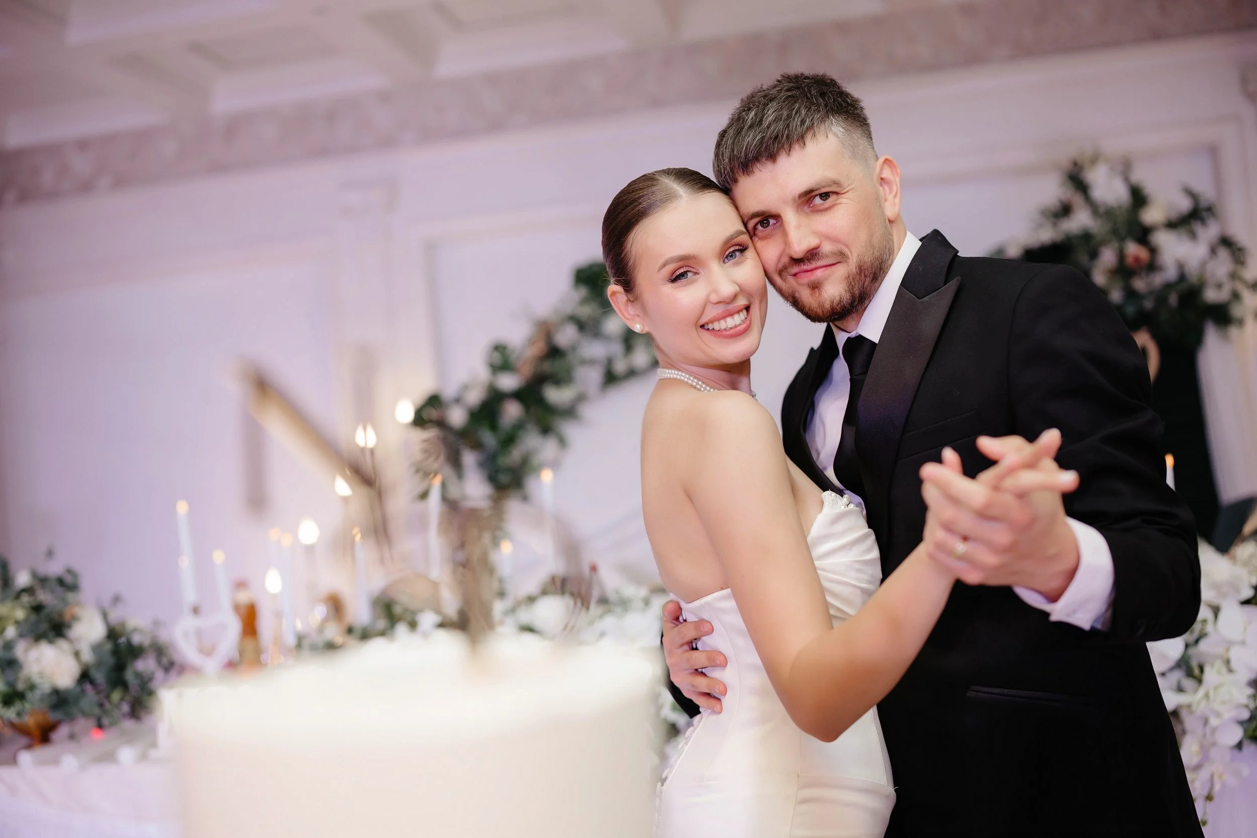 A newlywed couple is dancing at their wedding reception. The bride is wearing a white wedding dress and pearl jewelry, and the groom is dressed in a black tuxedo. They are smiling and embracing each other, with a decorated banquet hall with flowers a