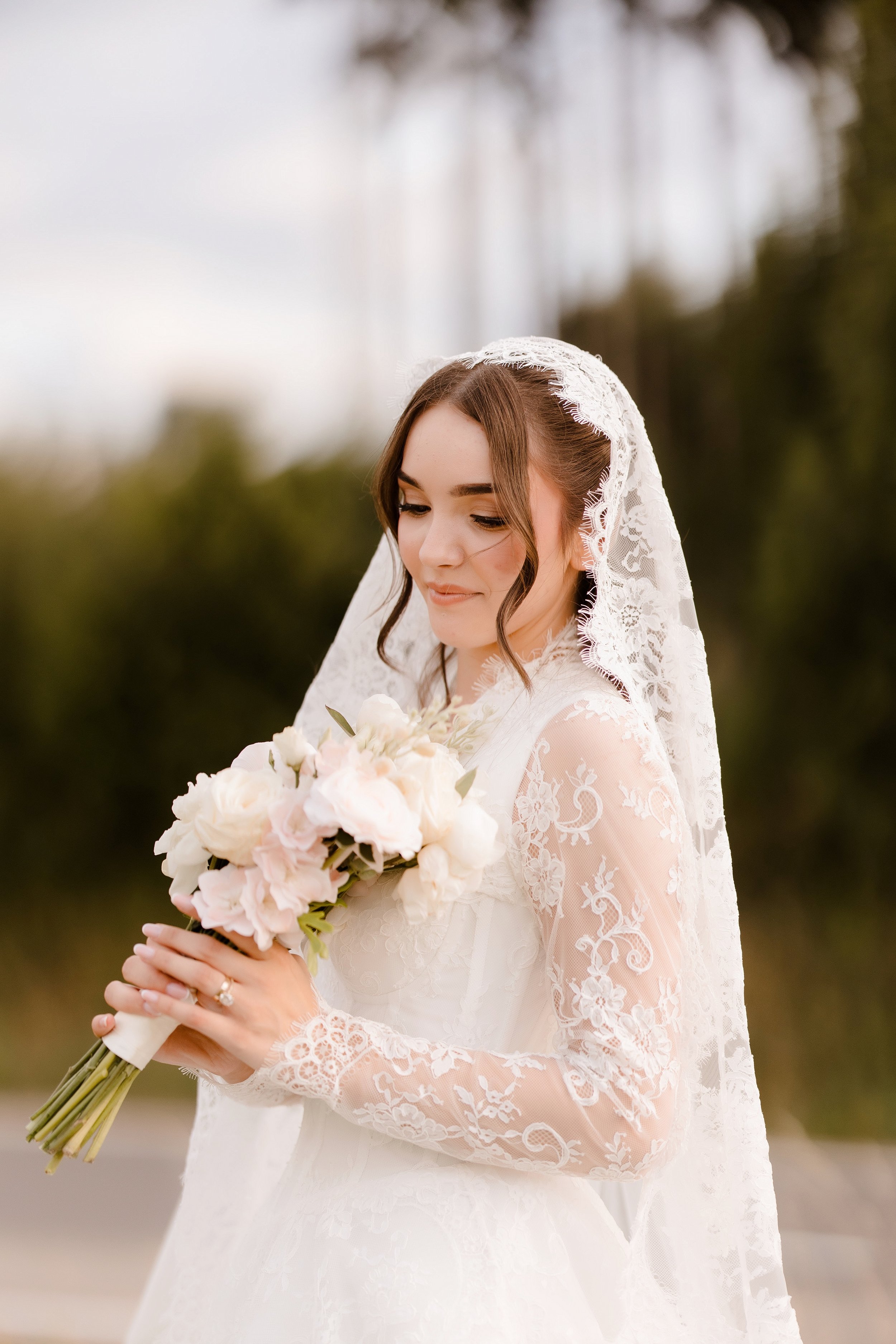 A bride in a white lace gown holding a bouquet of pink and white flowers, with a lace veil covering her head, outdoors.
