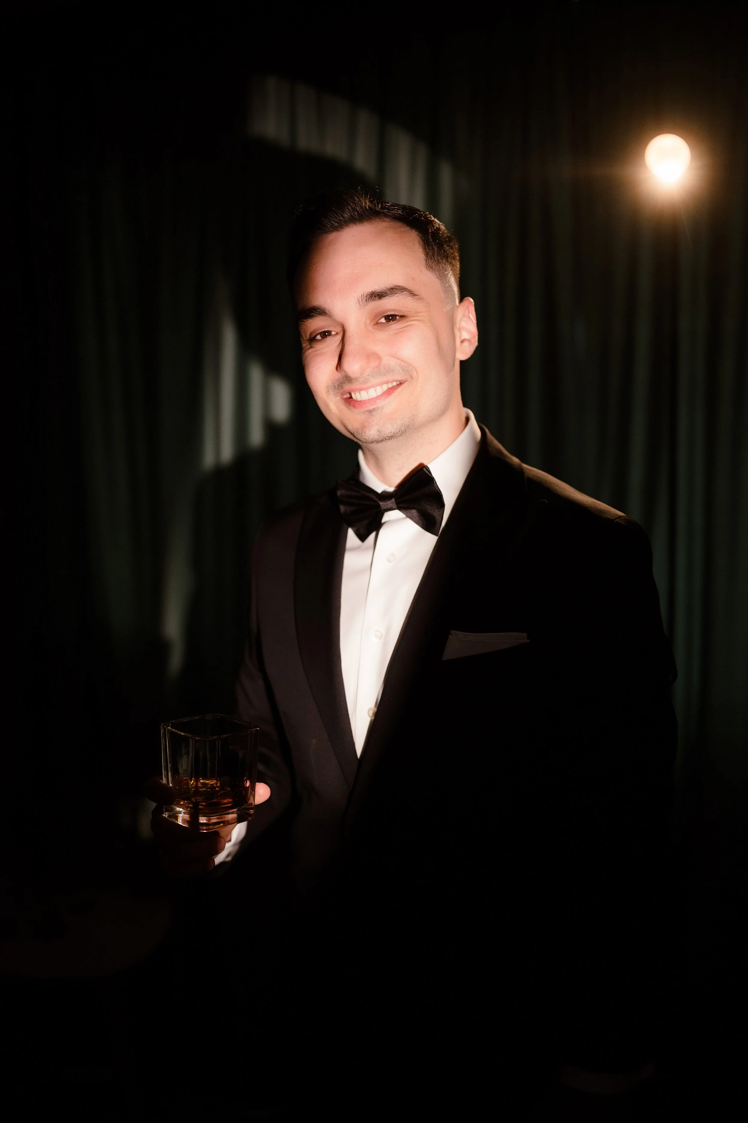 A man in a tuxedo holding a glass of a dark beverage, smiling warmly, in a dark setting with a bright, full moon in the background.