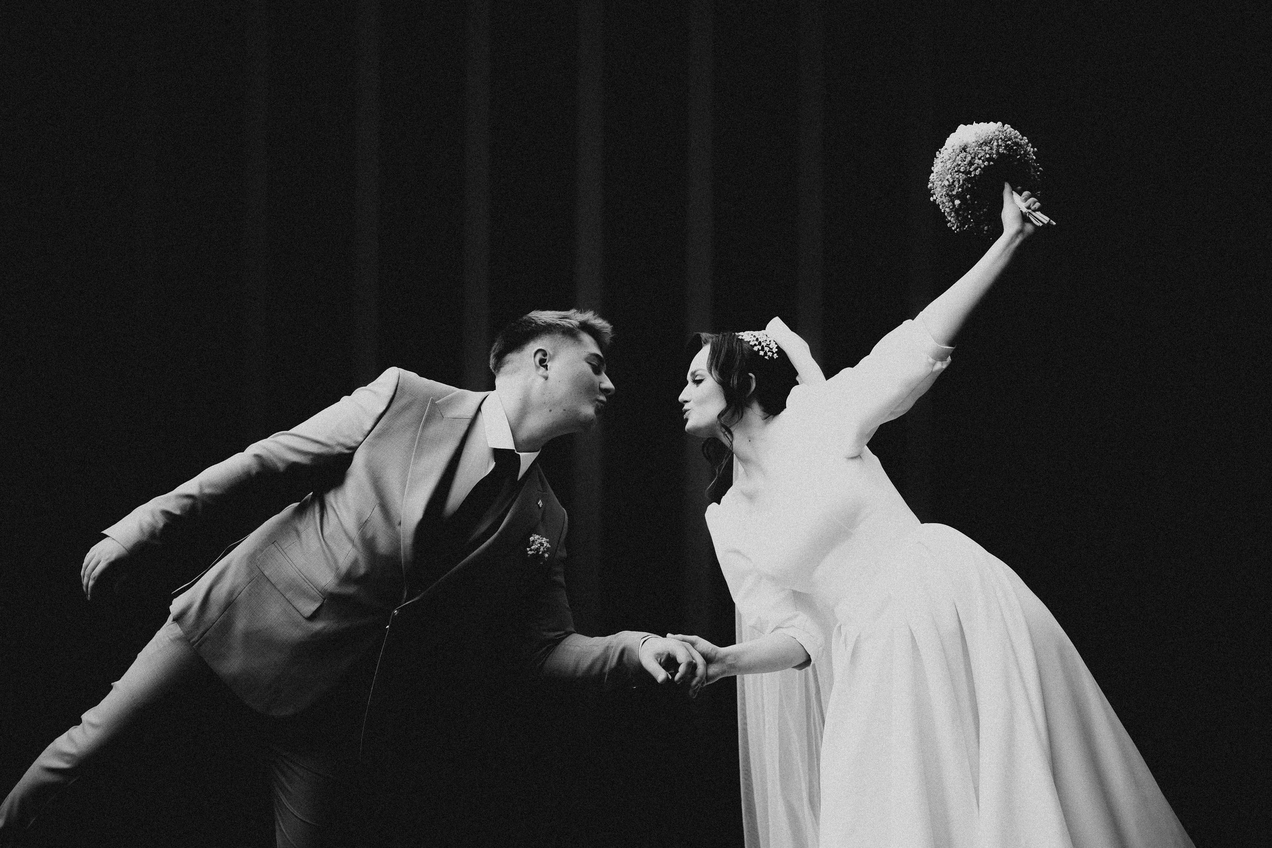 A black and white photo of a bride and groom leaning in close, holding hands, with the bride raising her arm holding a bouquet of flowers.