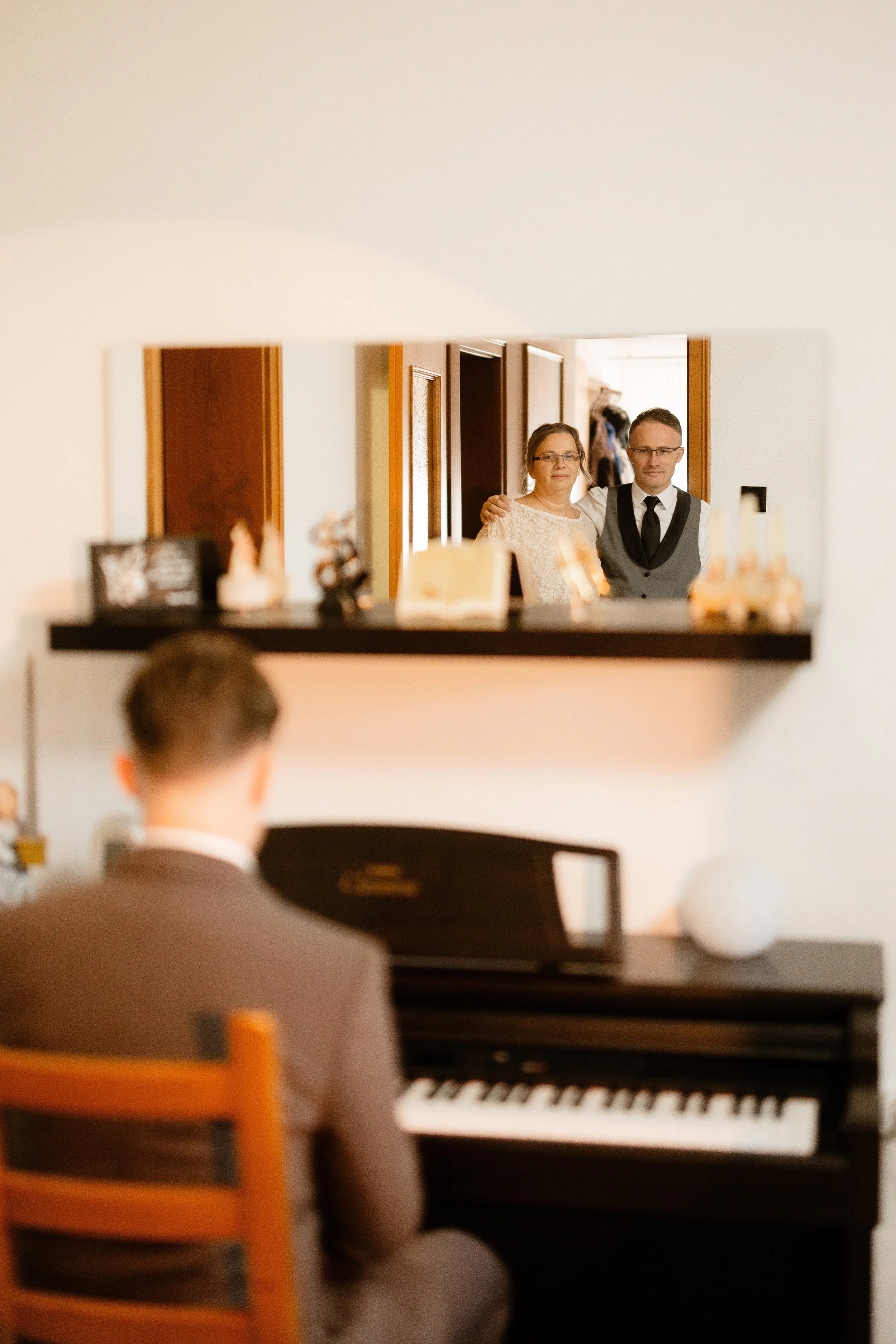 A woman and a man are seen in a reflection in a mirror, standing close together and smiling. The woman is wearing glasses and a white lace dress, while the man is dressed in formal attire with a vest and tie.