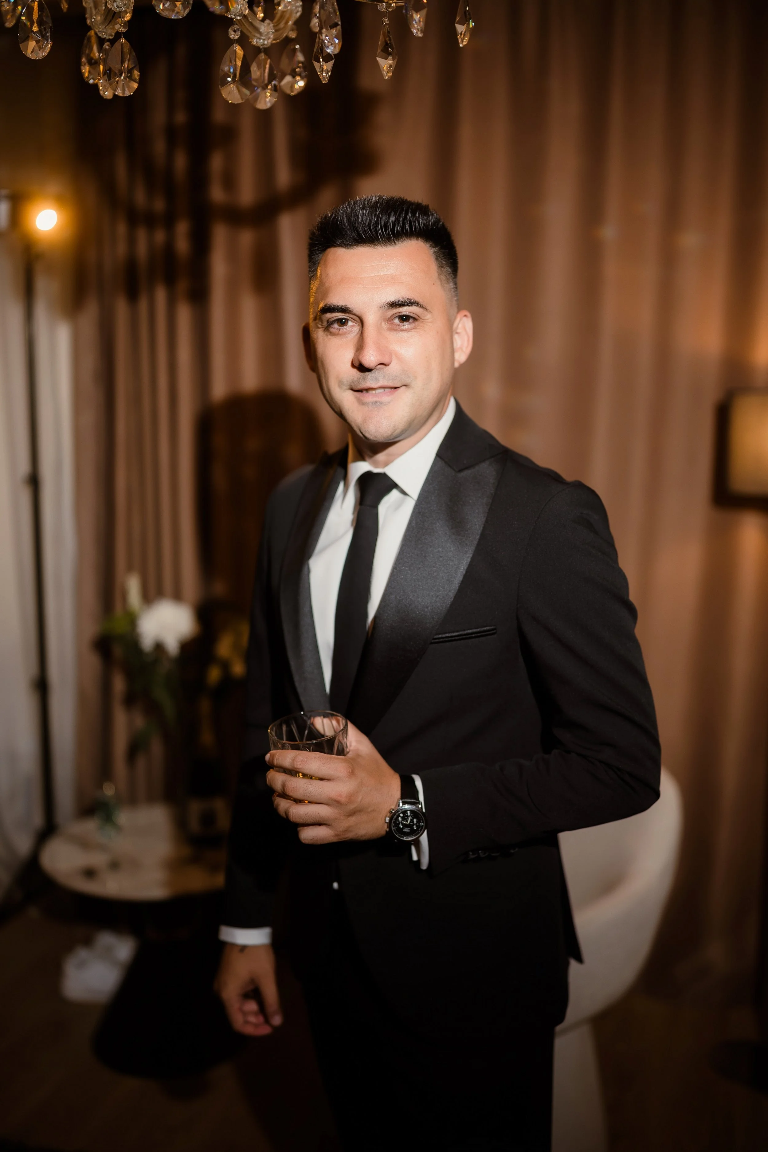 A man in a black tuxedo holding a glass of drink, standing in an indoor setting with elegant decor, including a chandelier and warm lighting.