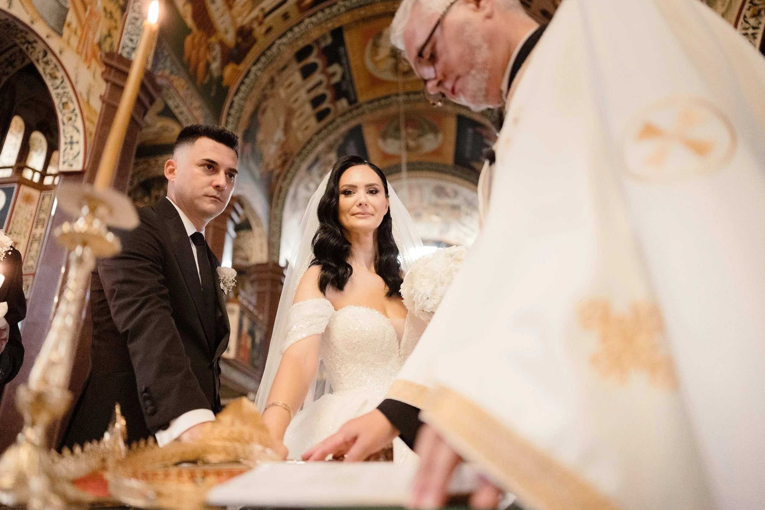 A bride and groom participating in a wedding ceremony inside a church, accompanied by a priest. The bride is wearing a white off-the-shoulder wedding dress and the groom is in a black suit. The priest is dressed in white robes and glasses, and they a