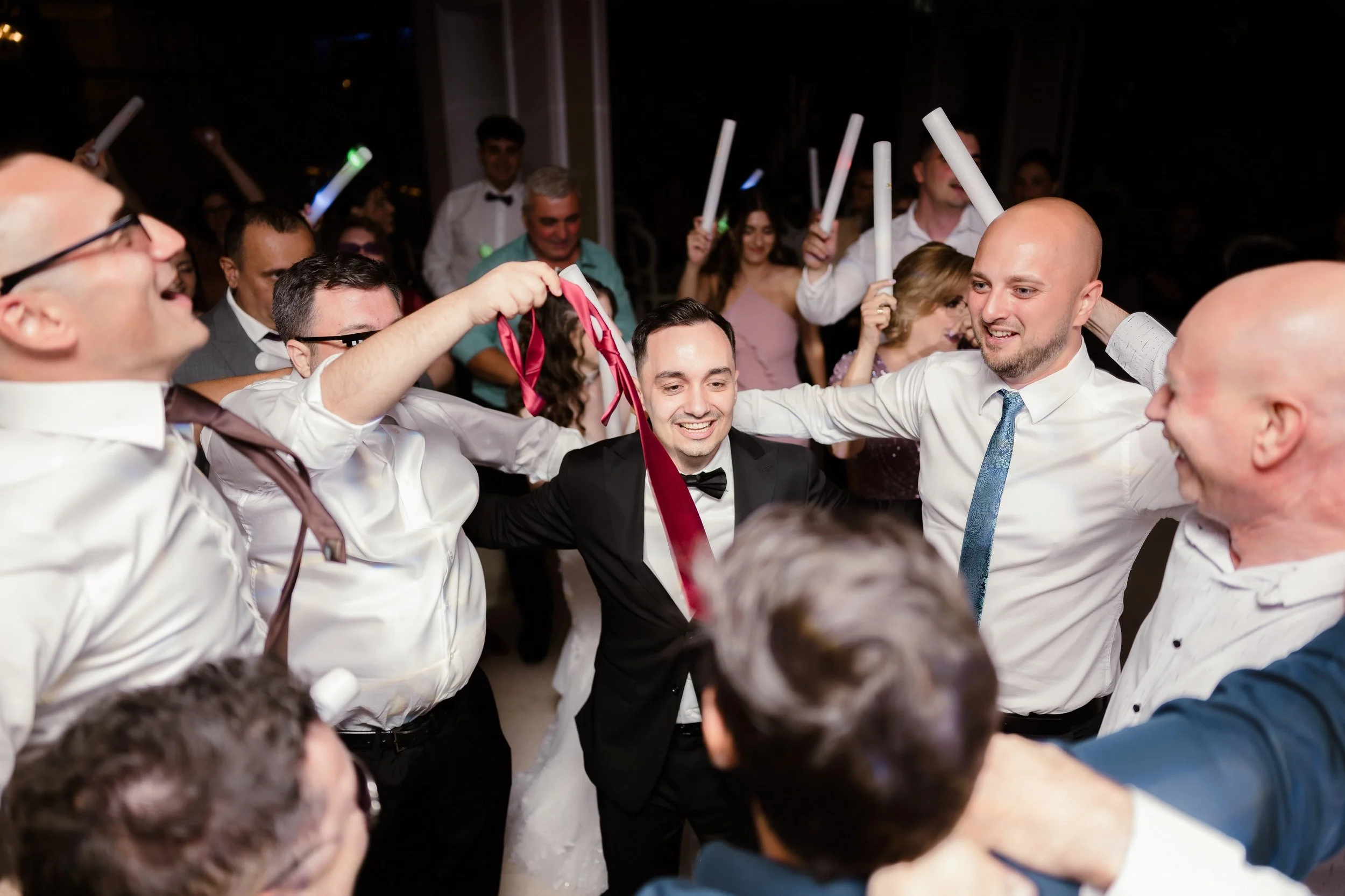 Group of people at a celebration, some holding glow sticks, with a man in a tuxedo at the center who appears to be the guest of honor.