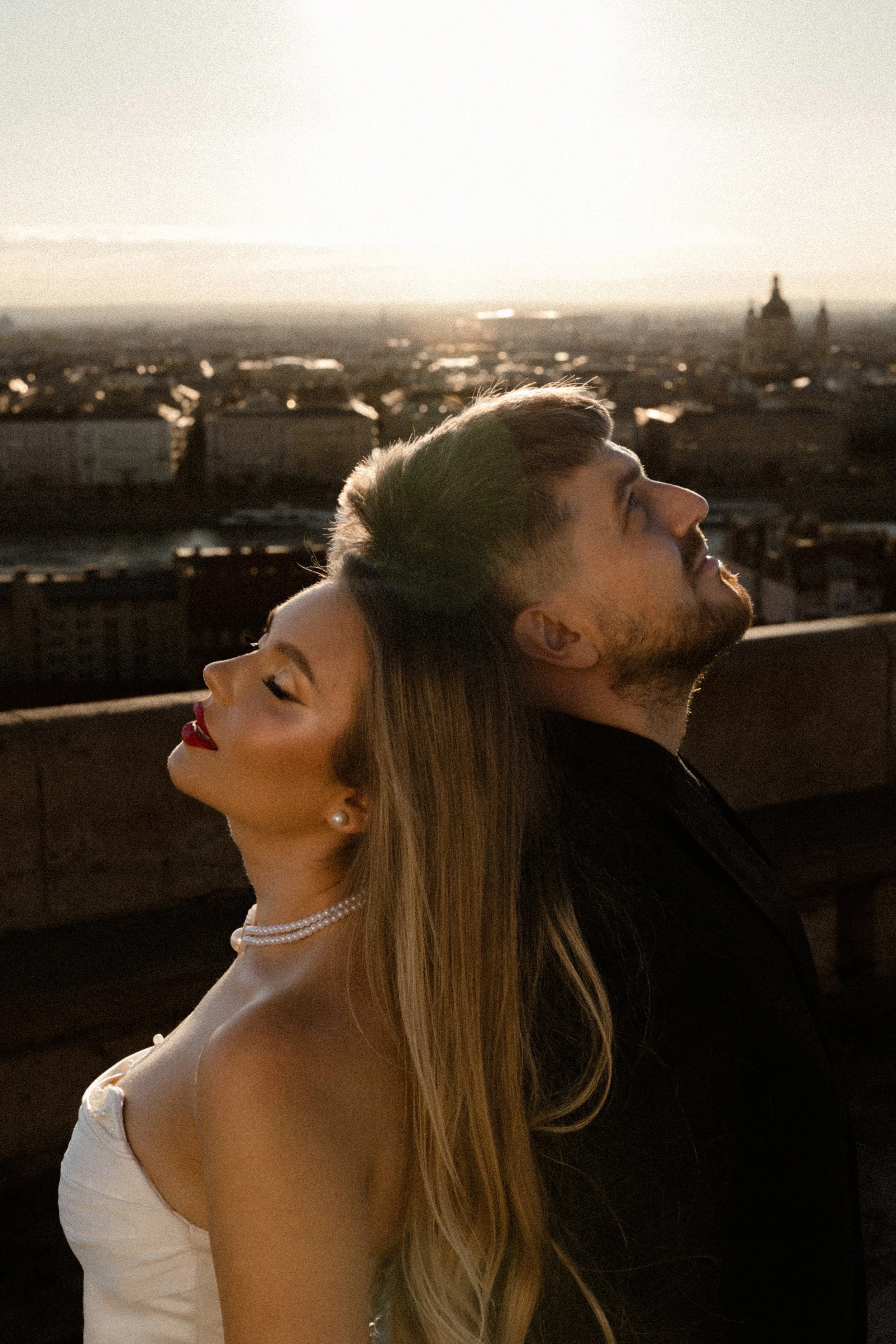 A man and a woman with their heads resting against each other, standing back to back with a cityscape and sunset in the background.