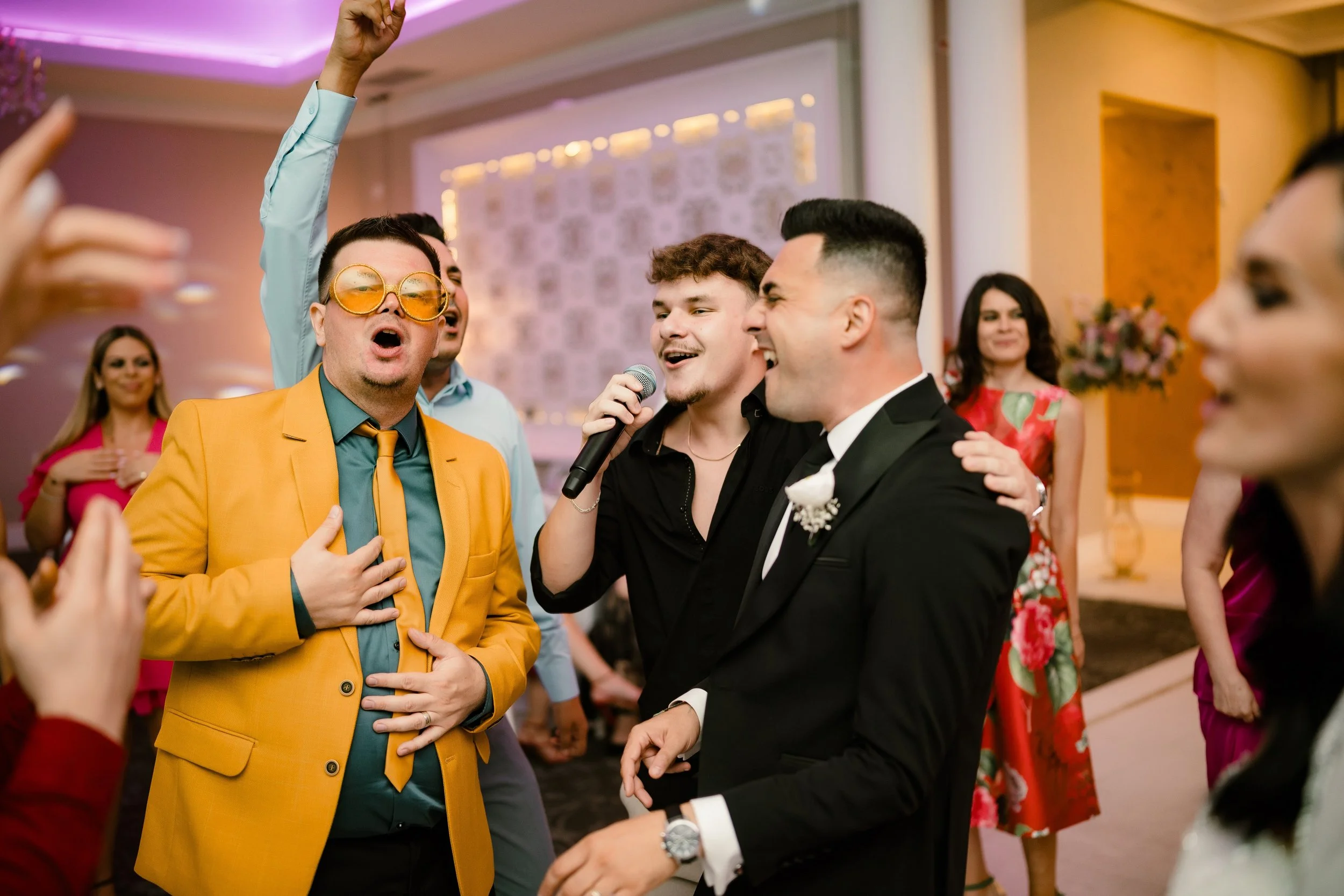 People celebrating at an event, some singing and dancing, in formal and colorful attire, with a woman in the background wearing a pink dress and women in floral dresses.