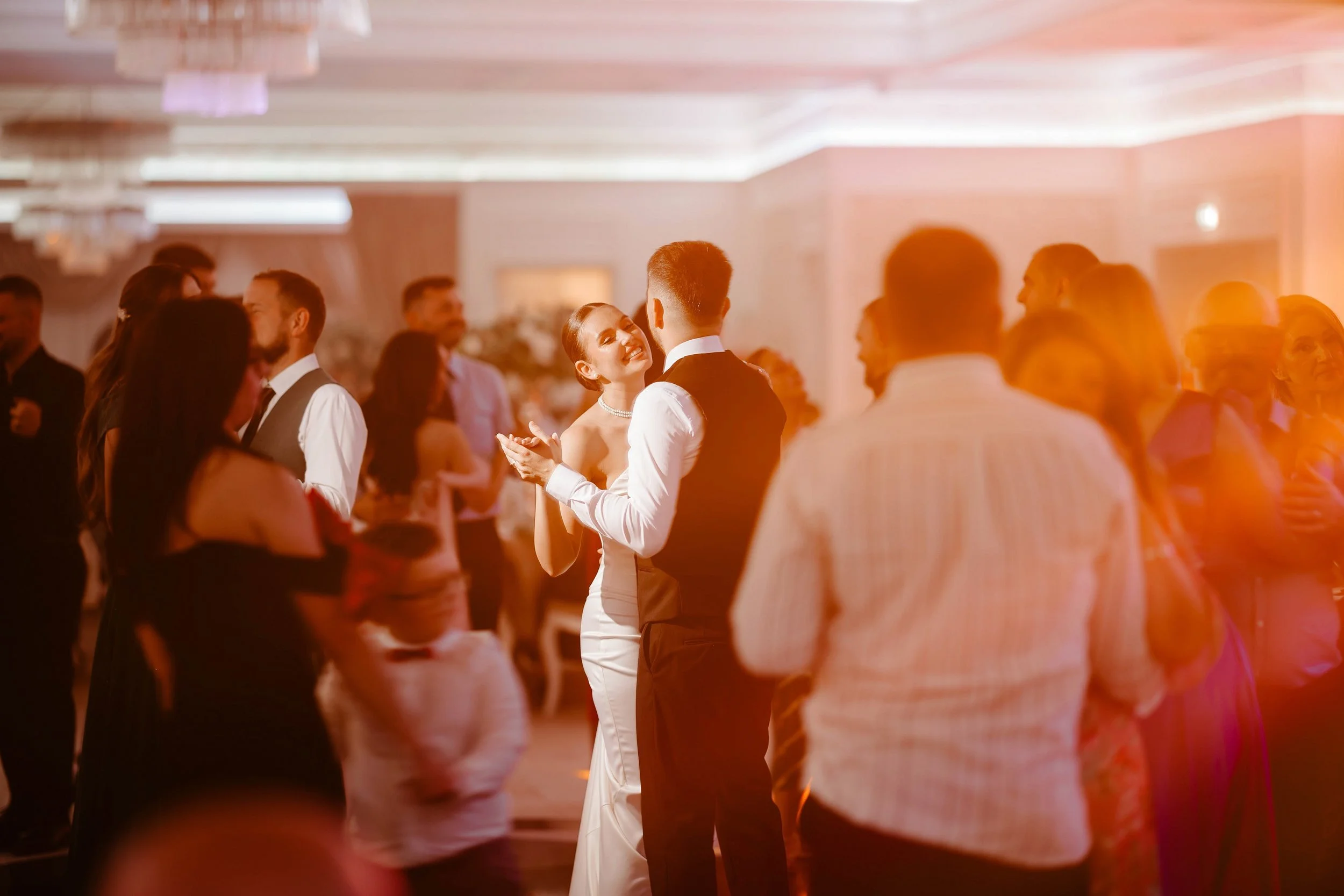 A couple dancing at a wedding reception with guests observing, in a warmly lit banquet hall.