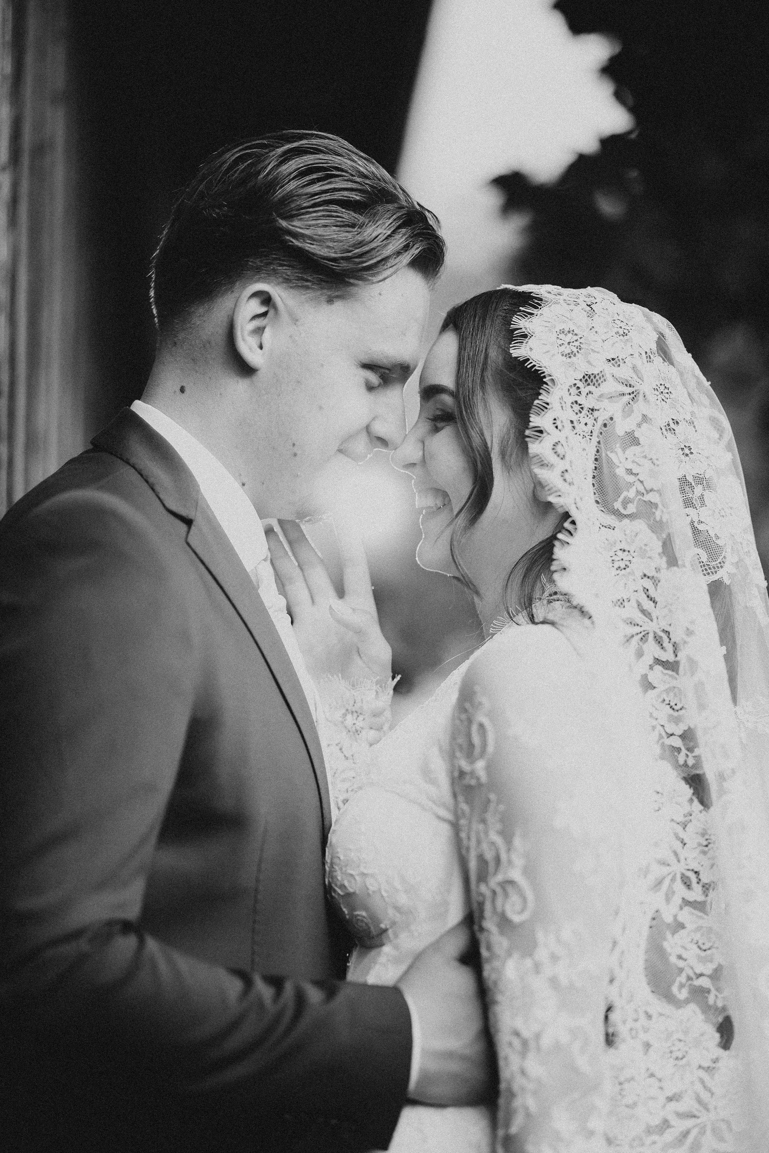 Black and white photo of a newlywed couple with their foreheads touching, smiling, with closed eyes. The bride wears an intricate lace wedding dress and a lace veil, while the groom is in a suit with slicked-back hair. The background is blurred, emph