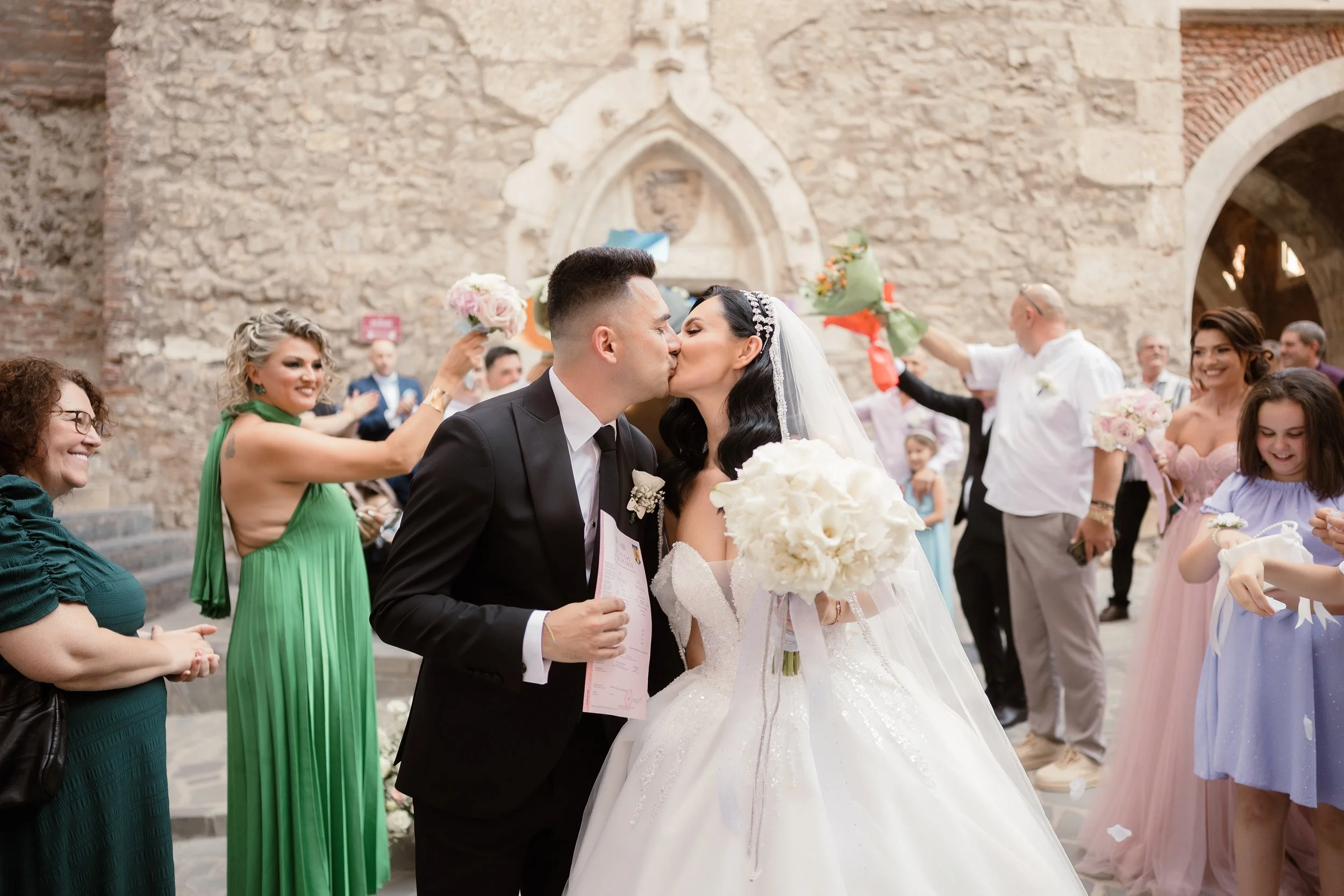 A bride and groom kiss during their wedding ceremony outside in front of a brick wall, surrounded by friends and family holding flowers and smiling.