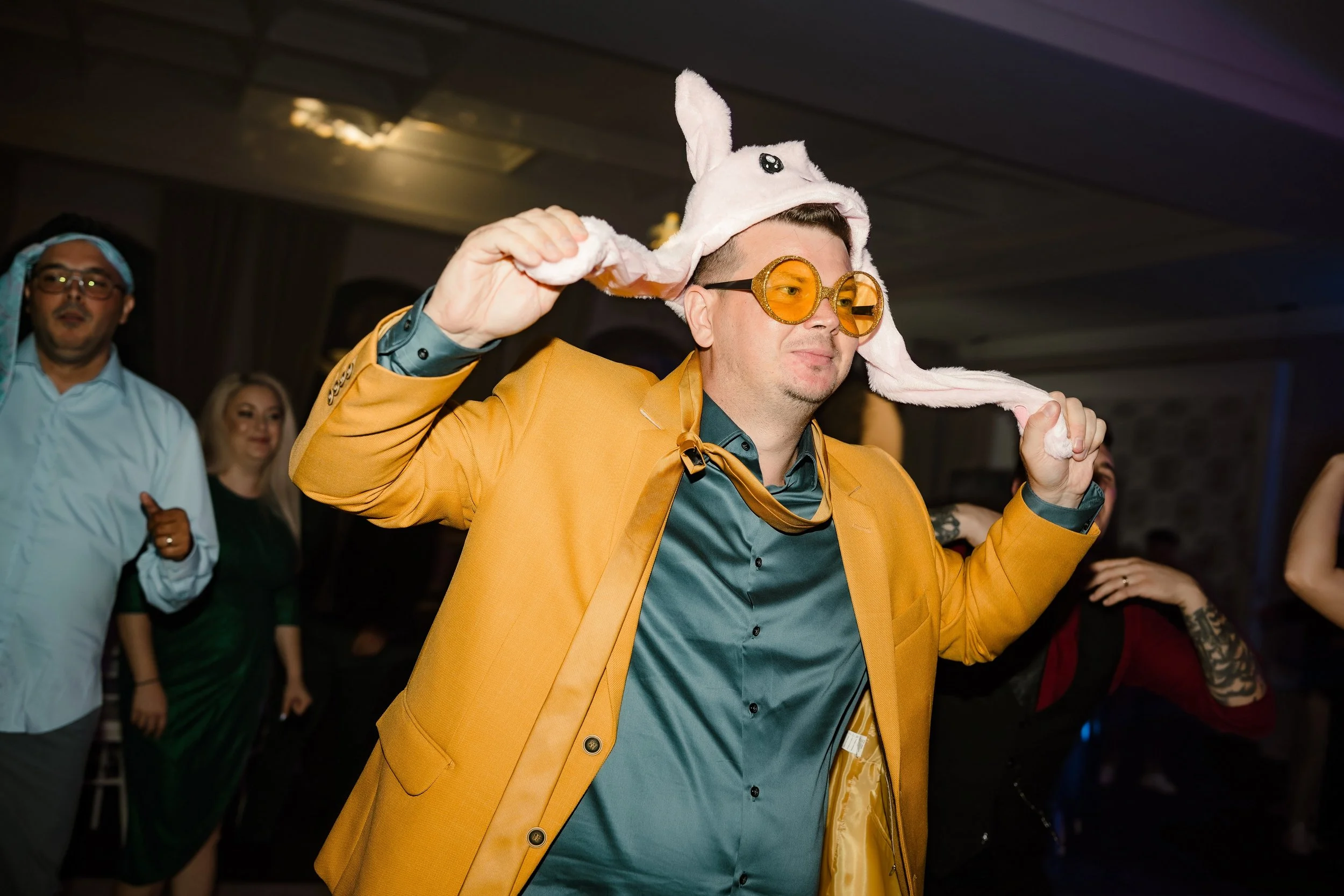 A man wearing a yellow jacket, turquoise shirt, and large yellow-tinted glasses, with a white bunny hat on his head. He is at a party or event, dancing or celebrating, with other people in the background.