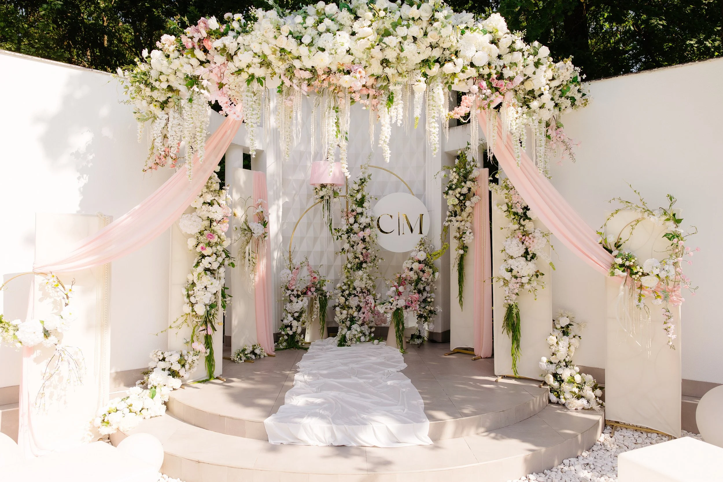 Elegant wedding altar decorated with cascading white and pink flowers, draped pink fabric, and a white circular sign with initials 'CM' on a white textured backdrop.
