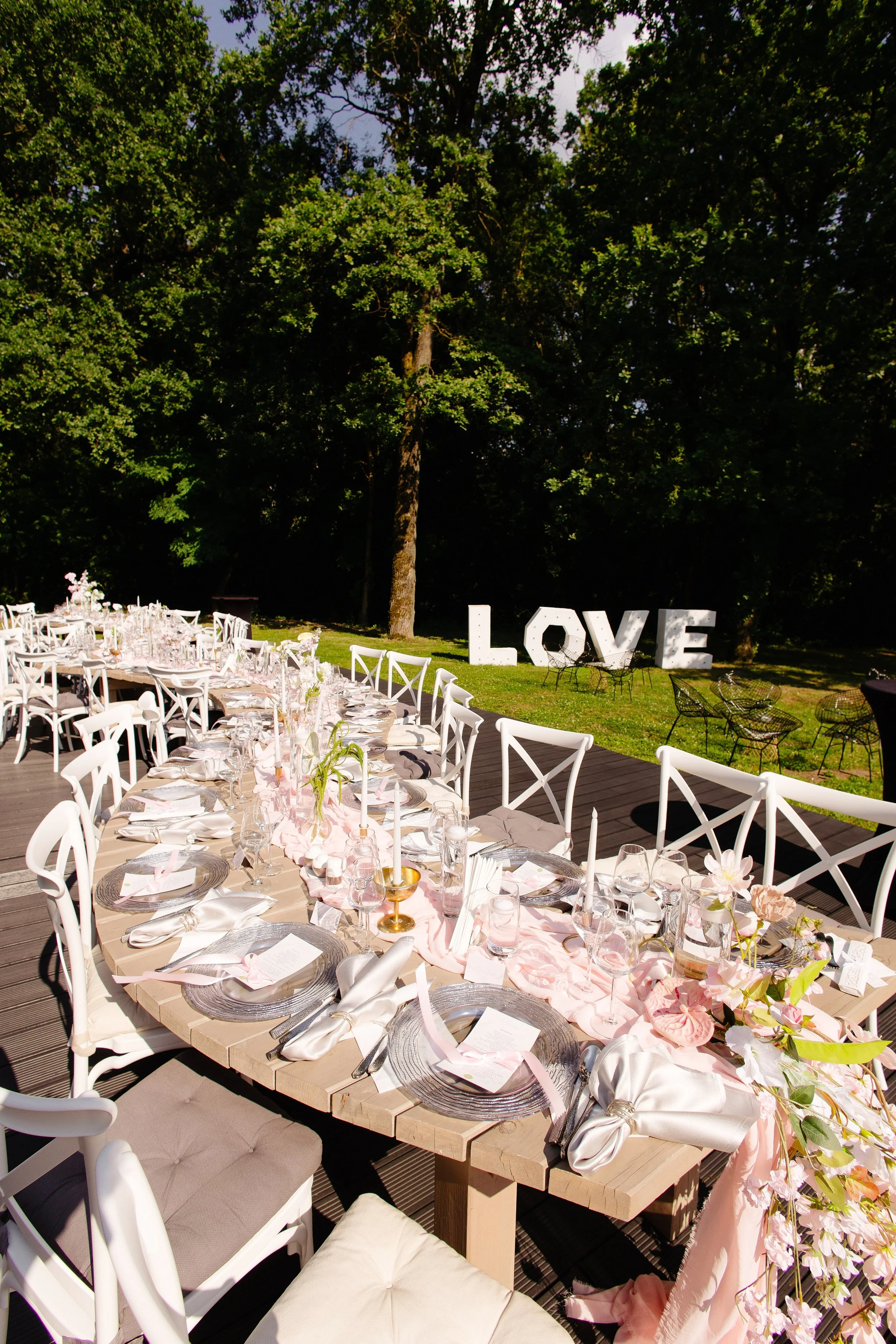 Outdoor wedding reception table setup with white chairs, pink table runner, floral decorations, and large white 'LOVE' sign in the background amidst lush green trees.
