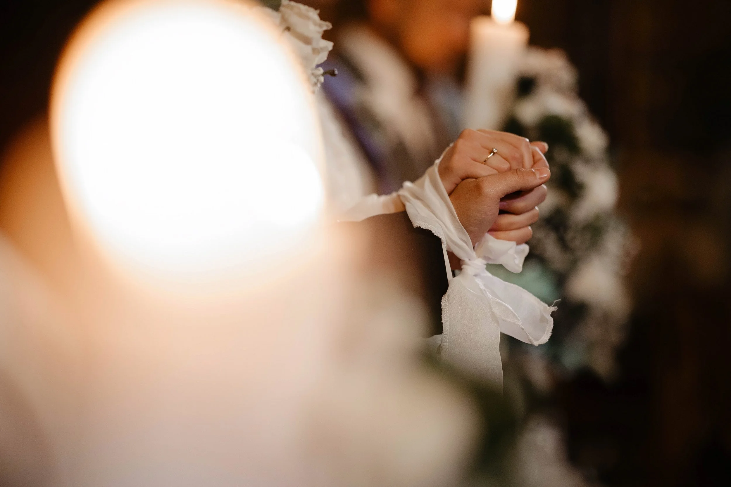 Close-up of hands clasped in prayer, with a wedding ring visible on the ring finger, in a dimly lit setting with candles in the background.