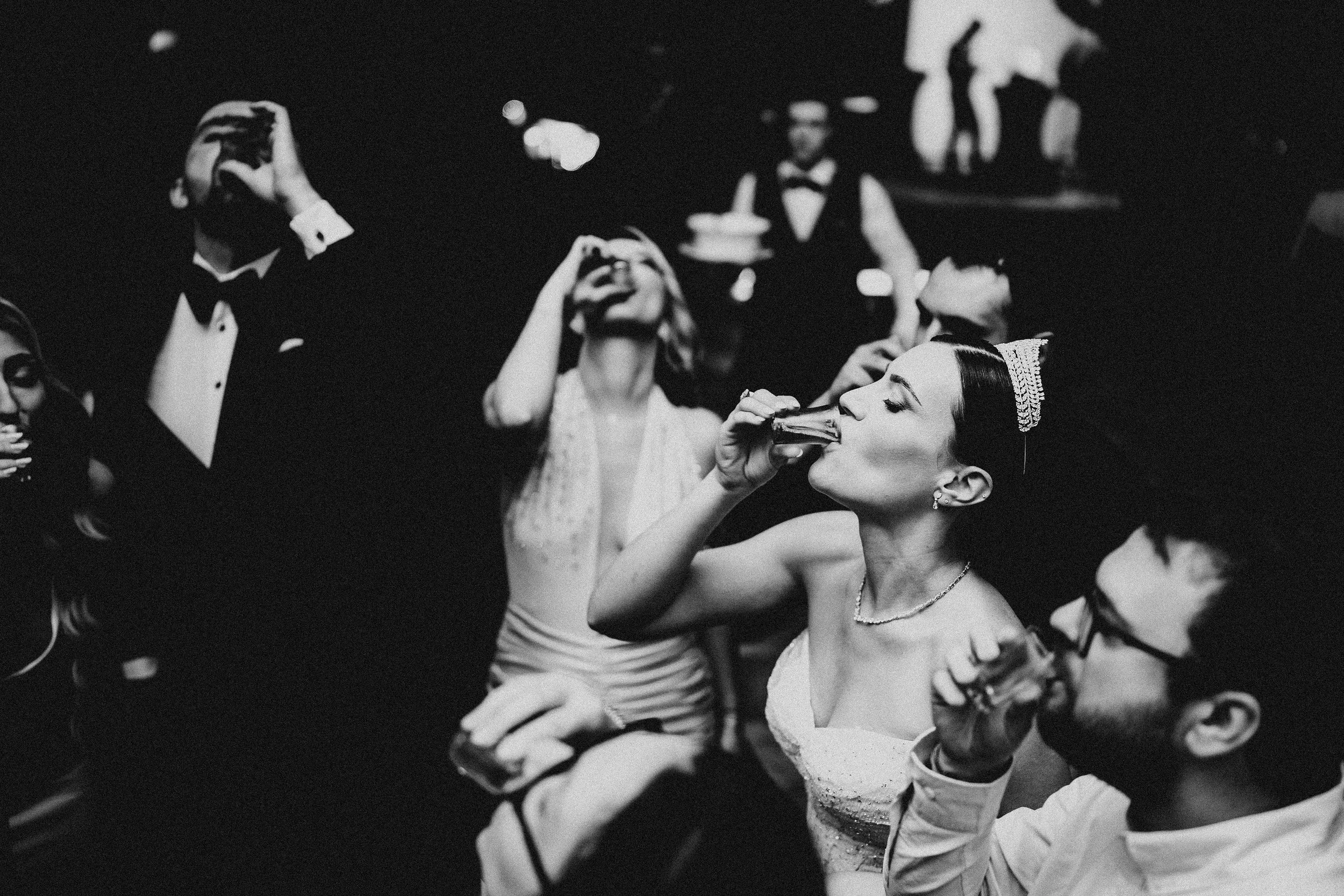 A group of elegantly dressed people at a formal event, with one woman drinking from a glass in the foreground and others socializing in the background.