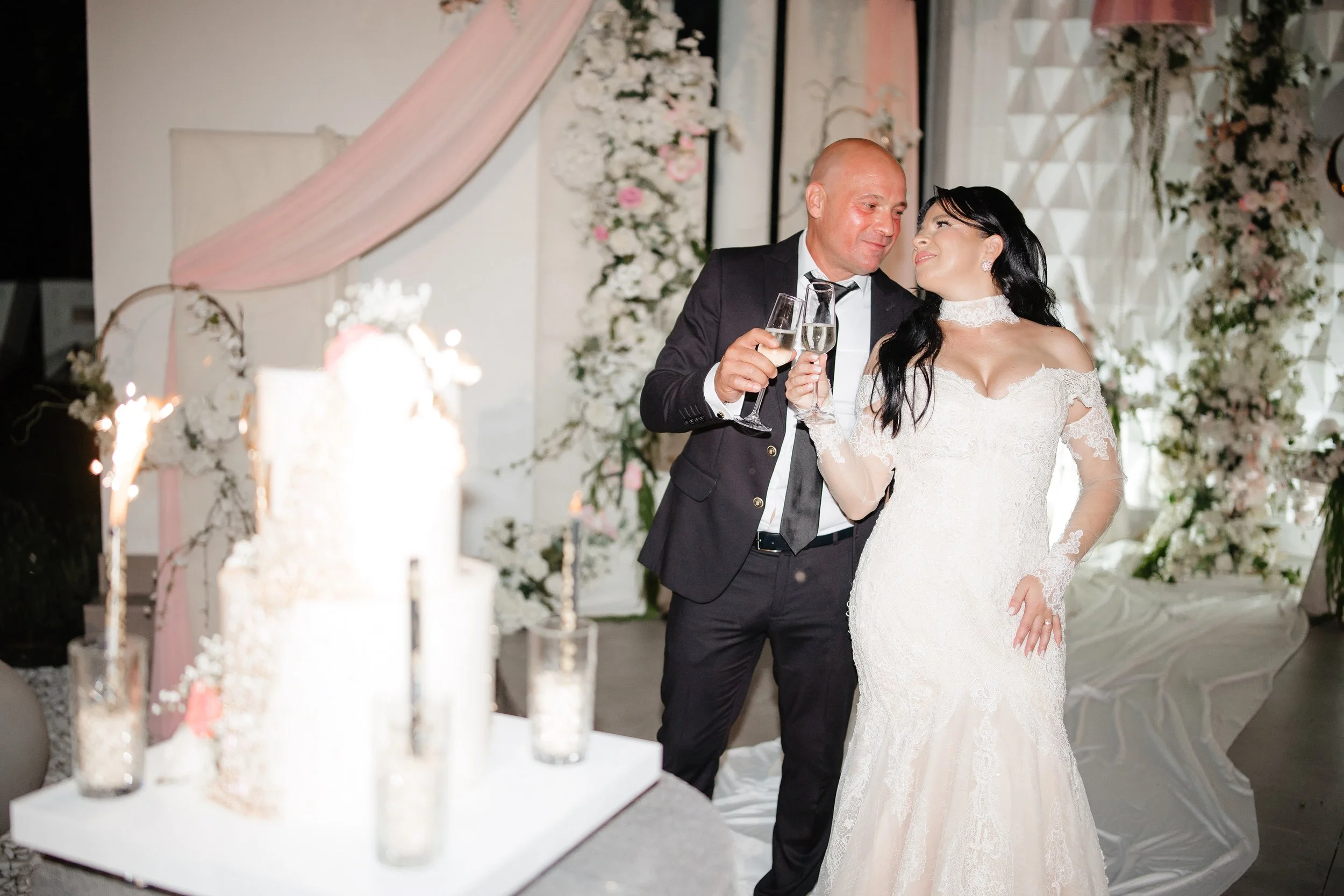 A bride and groom celebrating with champagne at their wedding reception. The bride is wearing a white lace wedding dress and the groom is in a black suit with a white shirt and black tie. They are smiling and looking at each other, holding champagne 