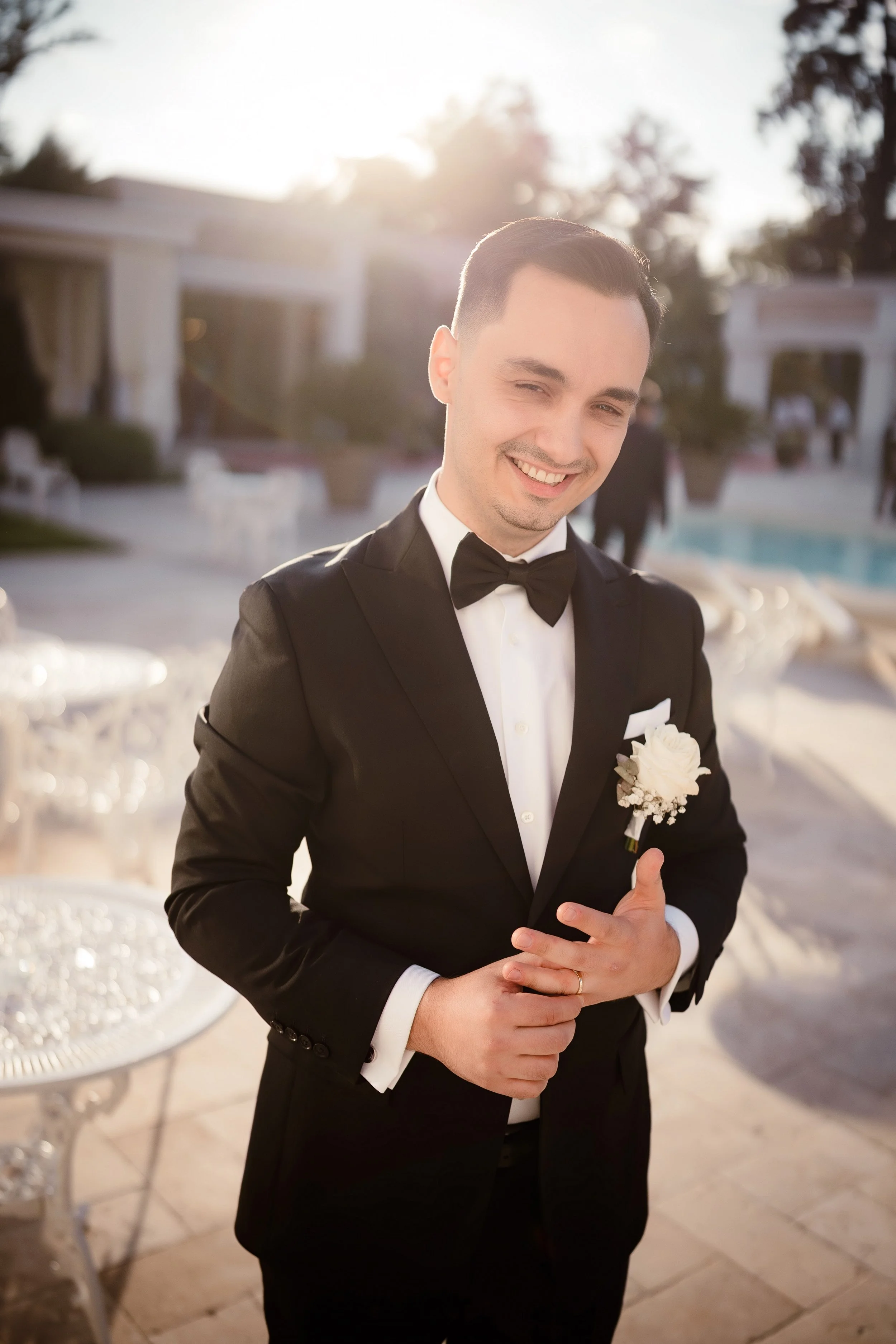 A young man dressed in a black tuxedo with a bow tie, smiling and giving a thumbs-up at an outdoor event during sunset.