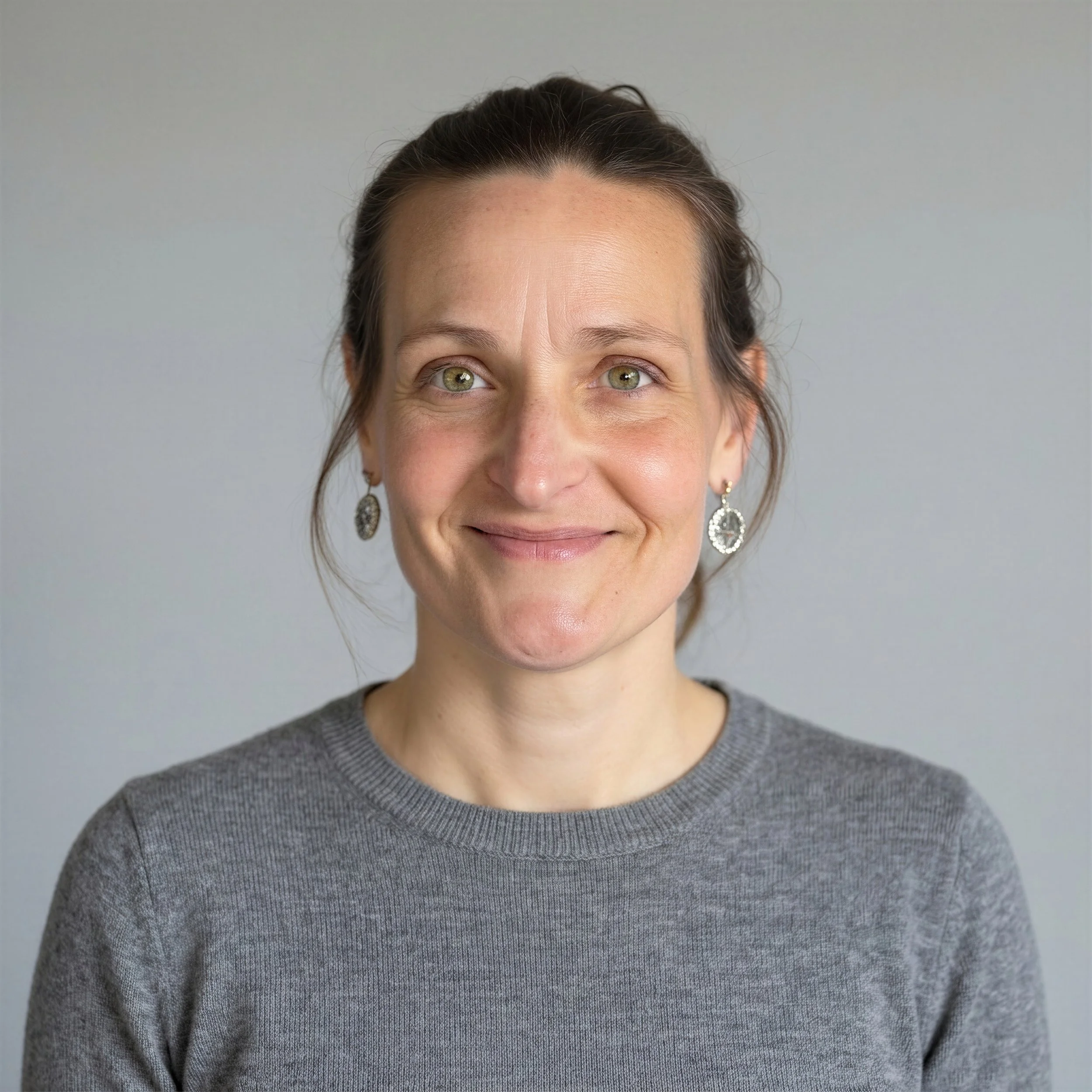 A woman with light skin and green eyes smiling, wearing a gray sweater and silver earrings, against a plain background.