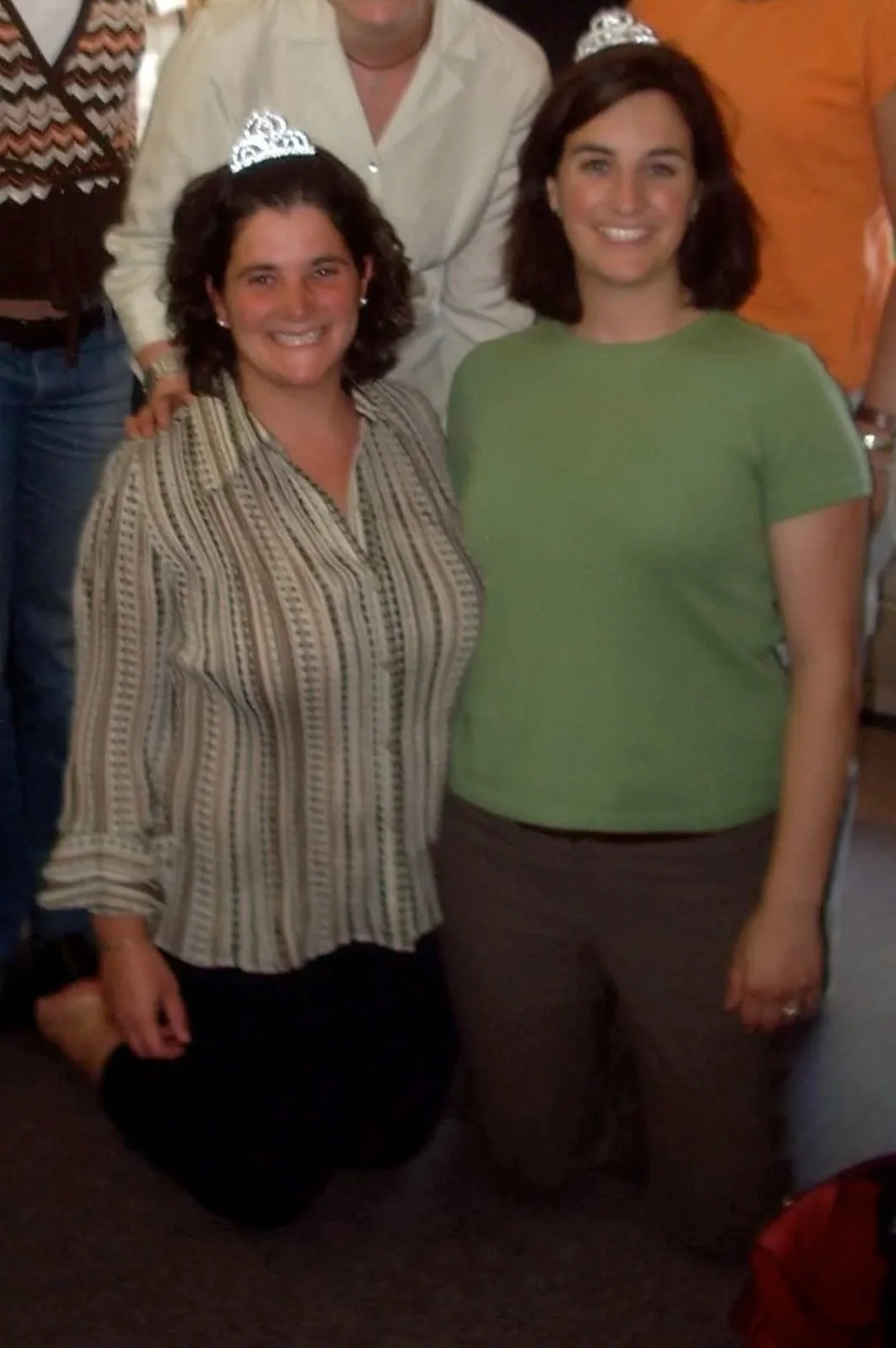 Two women wearing tiaras smile for a photo at a celebration. Both are dressed casually, and there are people standing behind them.