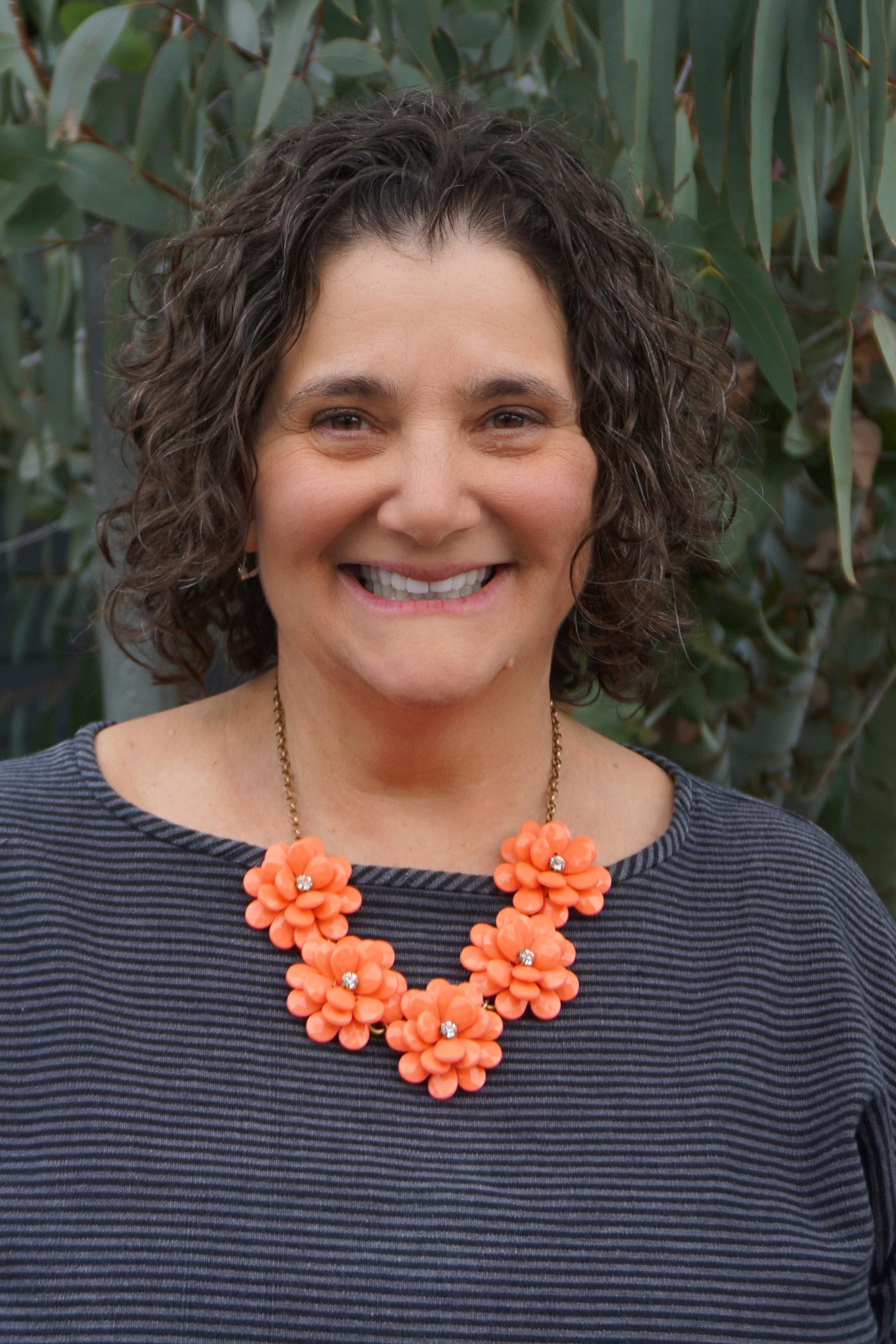 A woman named Laura Silverstein with short curly brown hair smiling and wearing an orange flower necklace and a dark striped top, standing in front of green plants.