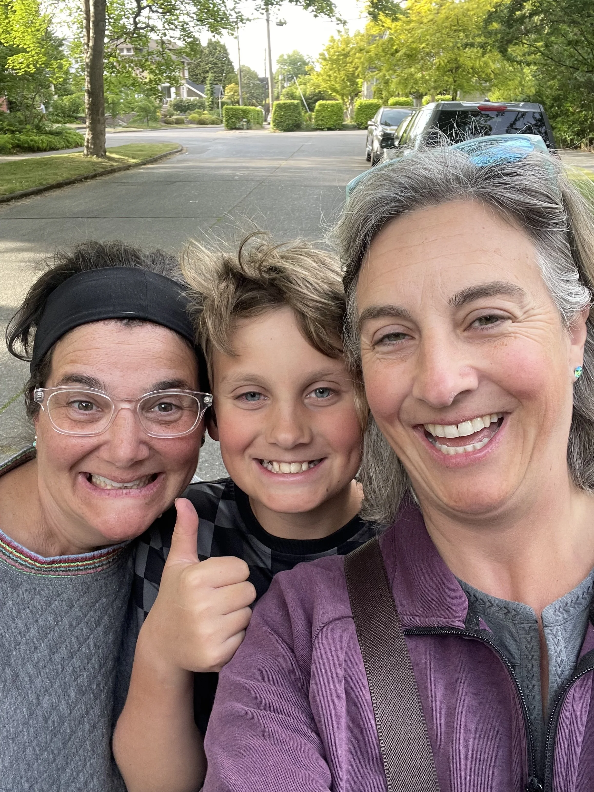 A group selfie of three smiling people, two women and a boy, outdoors on a sidewalk with trees, bushes, and parked cars in the background.