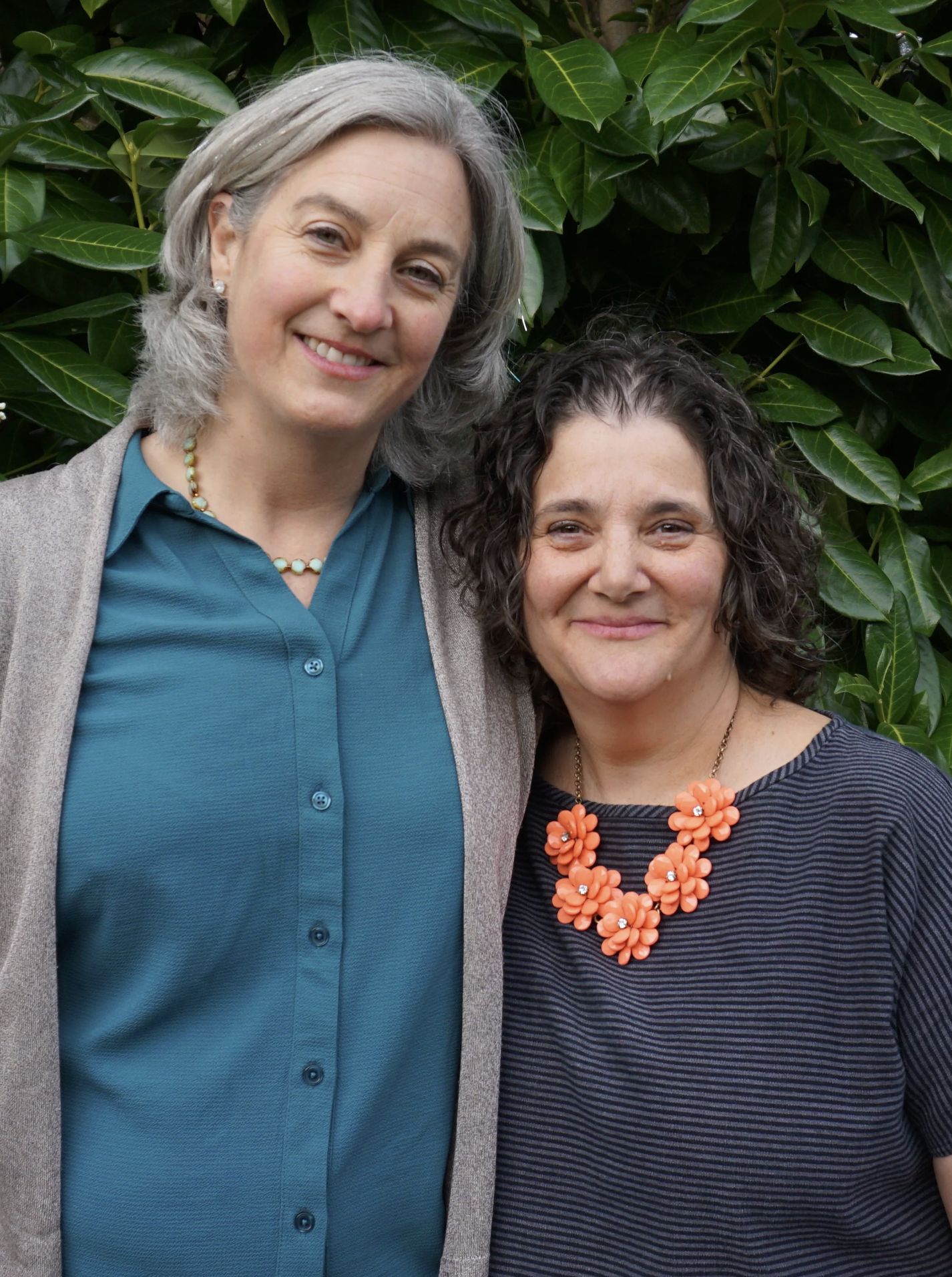 Two women, Sarah on left and Laura on right, smiling, standing close together in front of green leafy bushes.