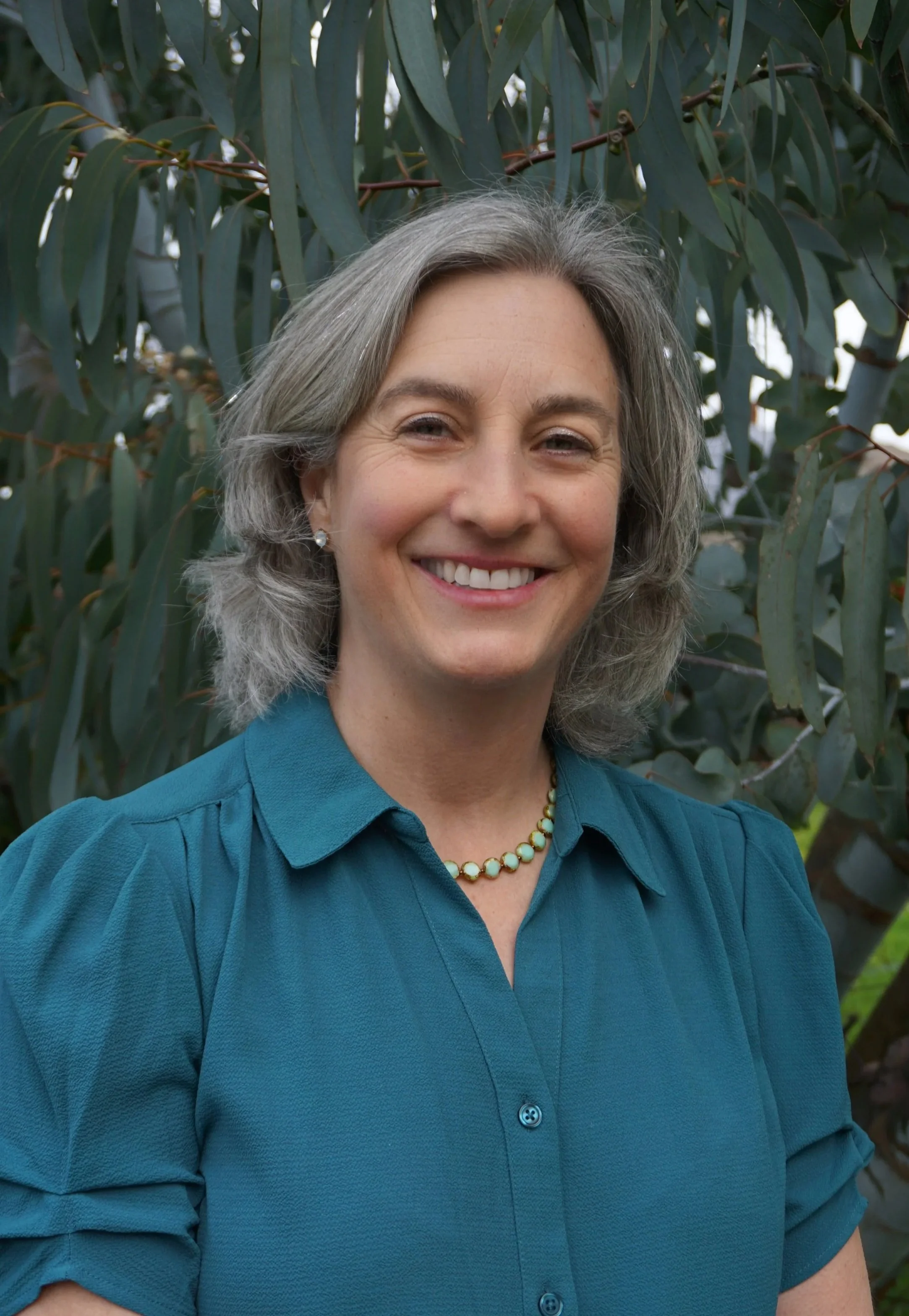A smiling woman named Sarah Schieron with gray hair wearing a teal shirt and pearl necklace standing in front of eucalyptus trees.