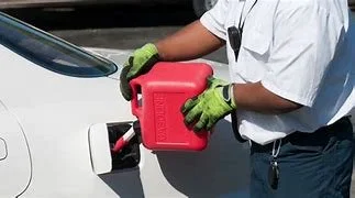 Person in white shirt and green gloves holding a red fuel can next to a white car.