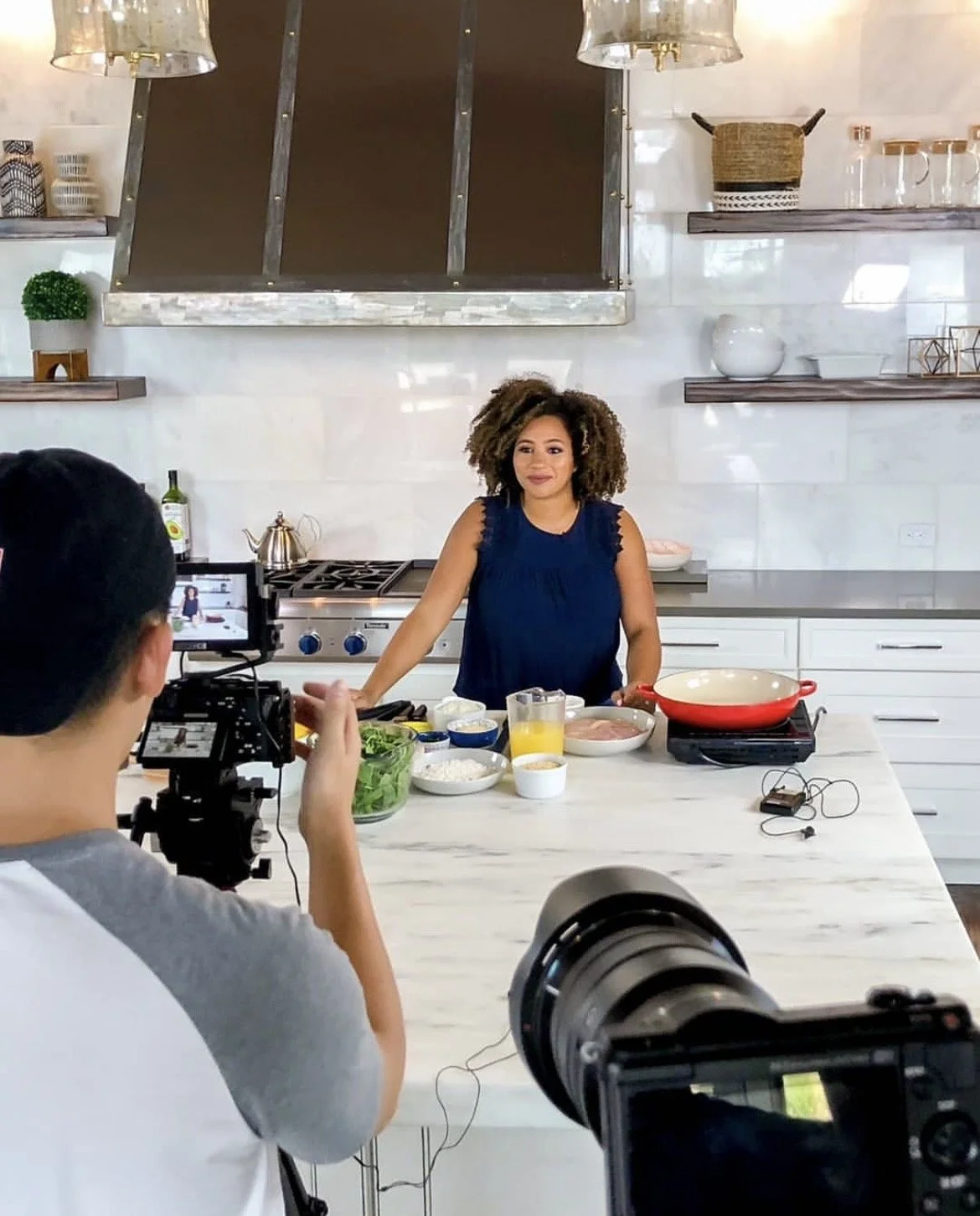 A woman with curly hair wearing a blue sleeveless top stands in a modern kitchen, preparing ingredients for a cooking demonstration. A man is filming her with a professional camera, capturing the scene. The kitchen has white cabinets, a marble countertop, and open shelves with decorative items.