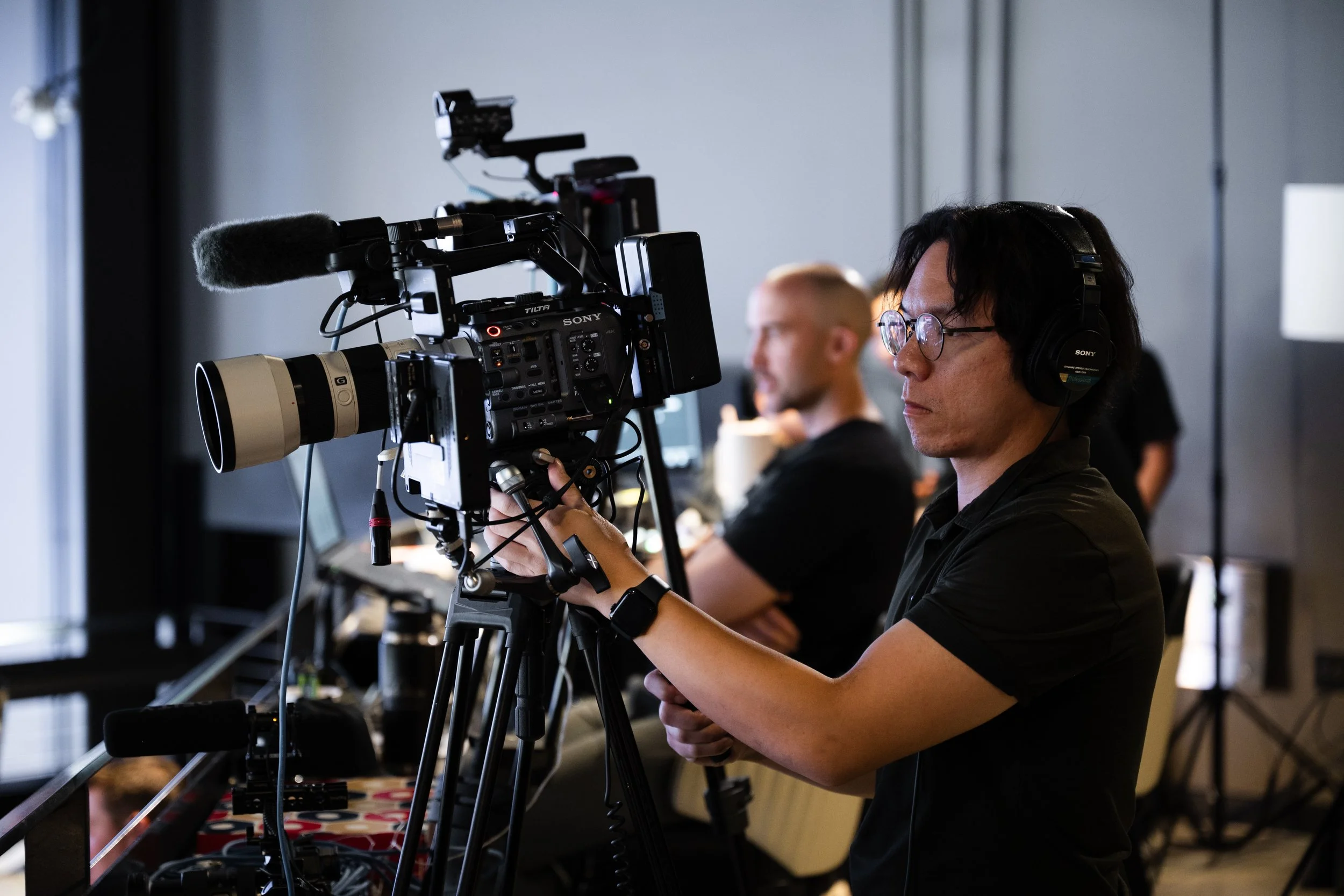 A person wearing glasses and headphones operating a professional video camera during a production or filming session, with other individuals blurred in the background.