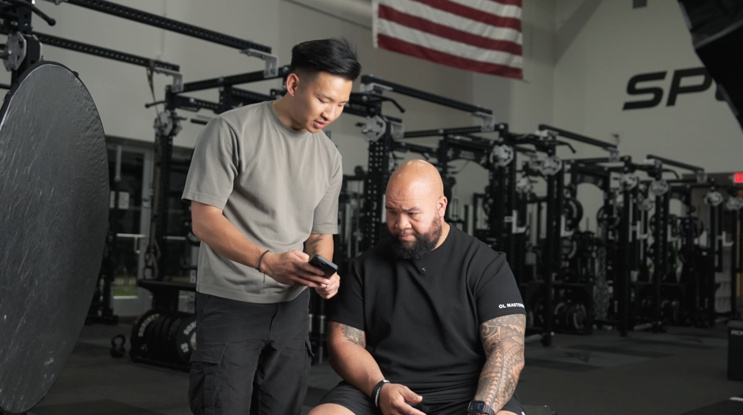 A trainer showing a phone to a man sitting on a bench in a gym with weightlifting equipment and an American flag in the background.