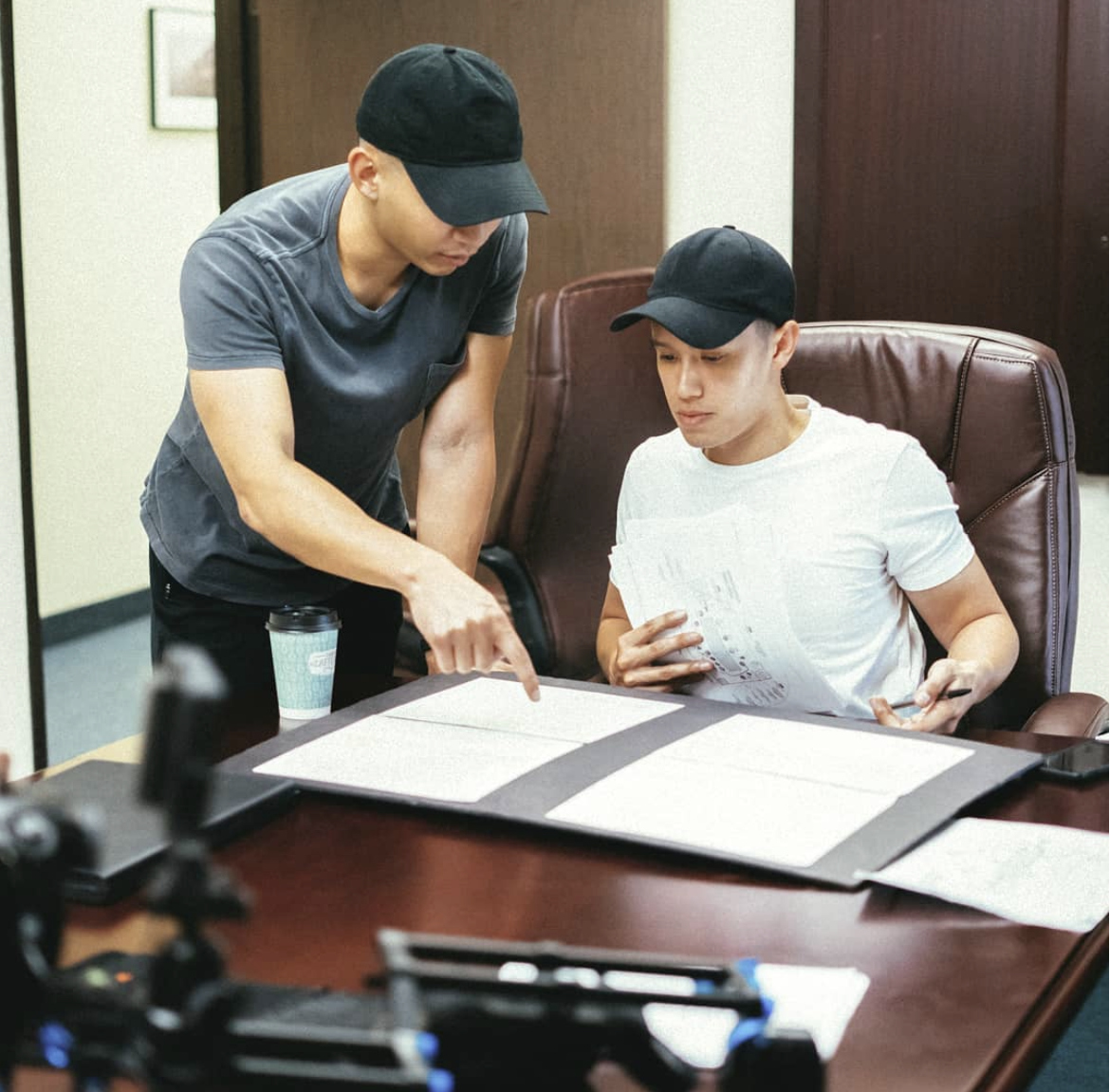 Two men wearing black caps and casual t-shirts are discussing documents on a conference table in a meeting room.