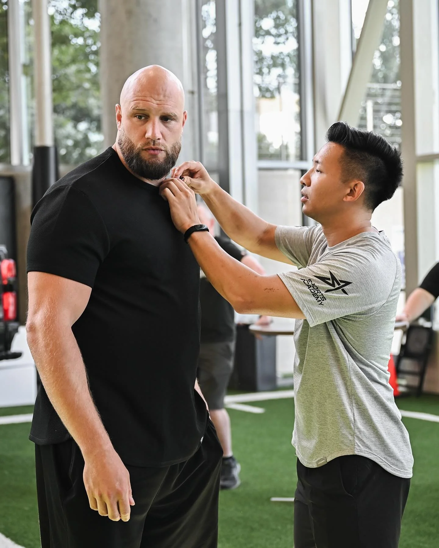 A man with a beard and a shaved head standing in a gym while a young man adjusts his collar.